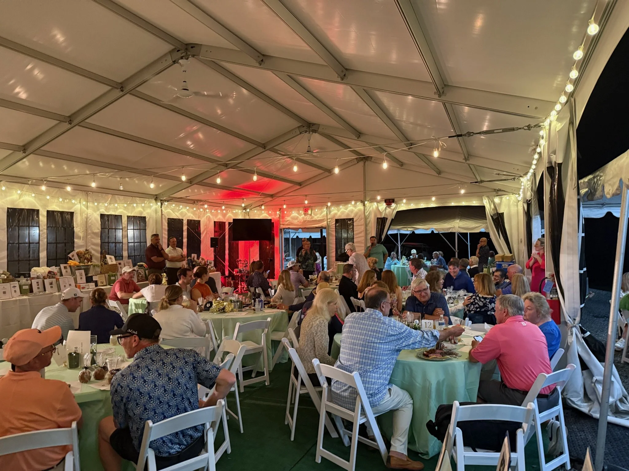 People sitting at tables under a large tent with string lights, during an evening event.