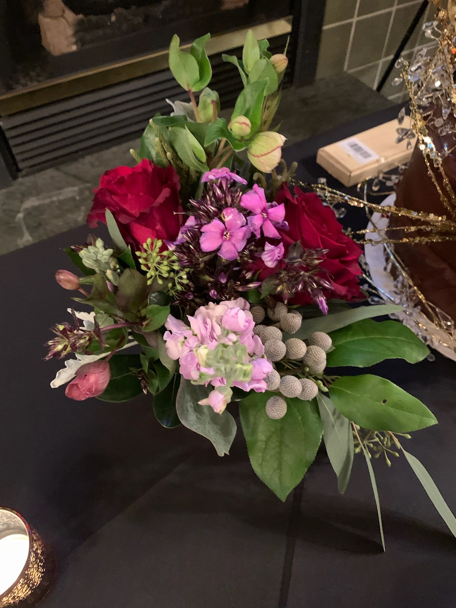 A floral arrangement with pink, red, and light purple flowers, surrounded by green leaves, on a black surface with a black fireplace in the background.