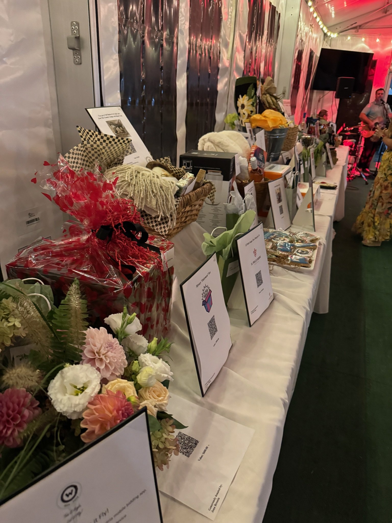 A display table with various items including flowers, baskets, and informational signs in a tent with string lights, with musicians performing in the background