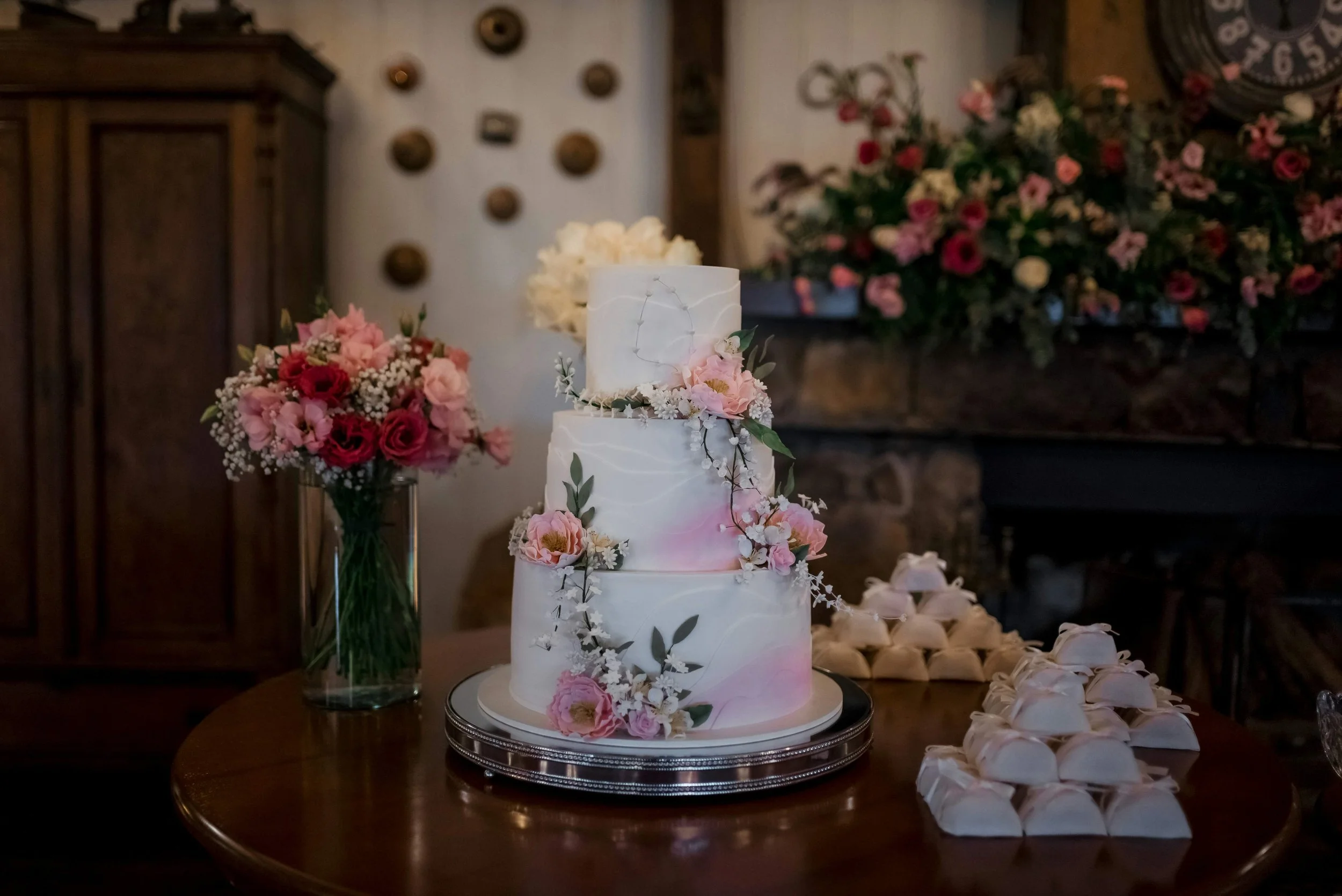A multi-tiered white wedding cake decorated with pink and white flowers, placed on a silver cake stand on a wooden table. Behind the cake, there are floral arrangements and a rustic background with a clock and wall decor.