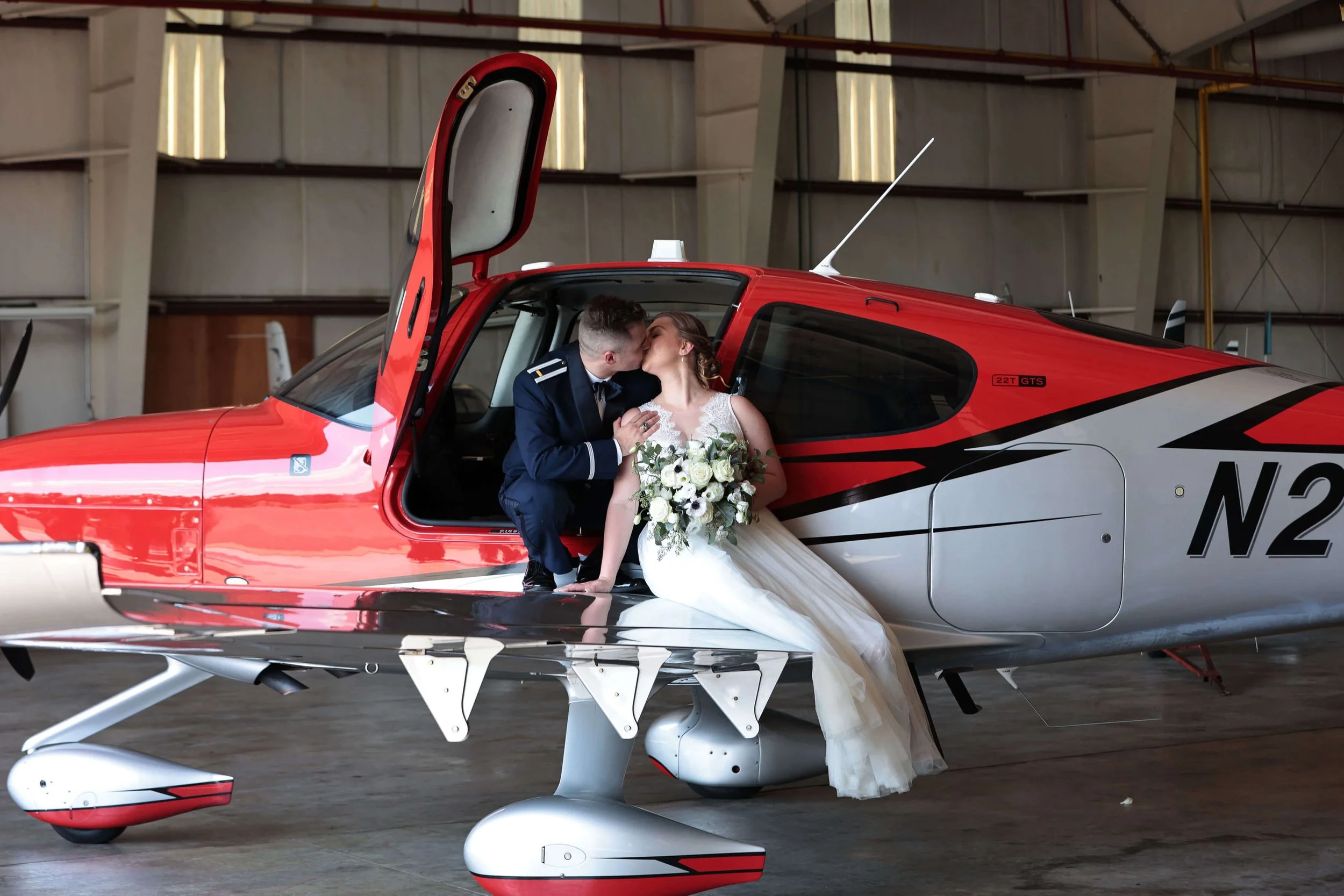 A newlywed couple kissing inside a red and white small aircraft in an airplane hangar, with the bride holding a bouquet of white flowers.