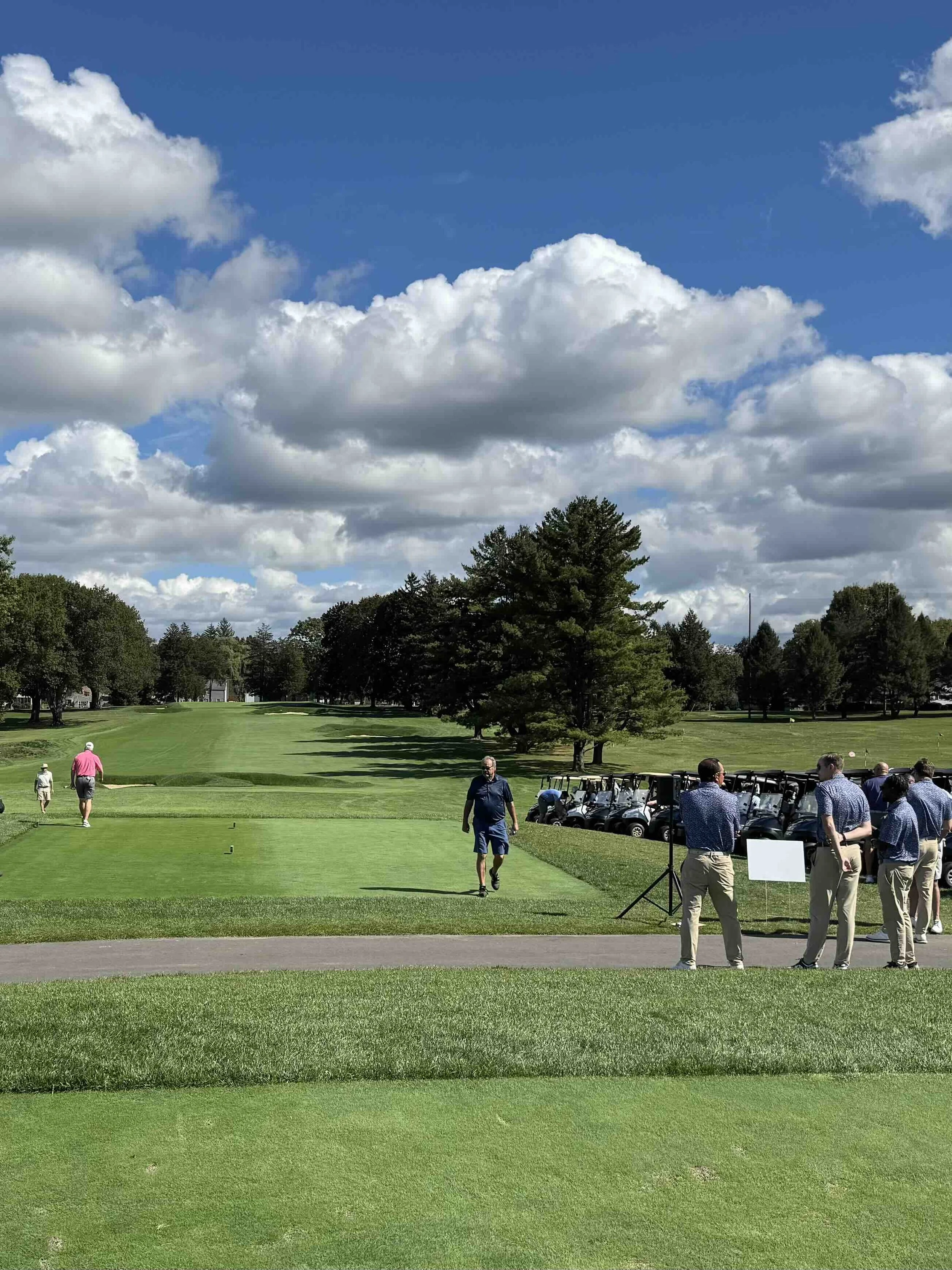 A group of people playing golf on a lush green golf course on a sunny day with partly cloudy skies and trees in the background.