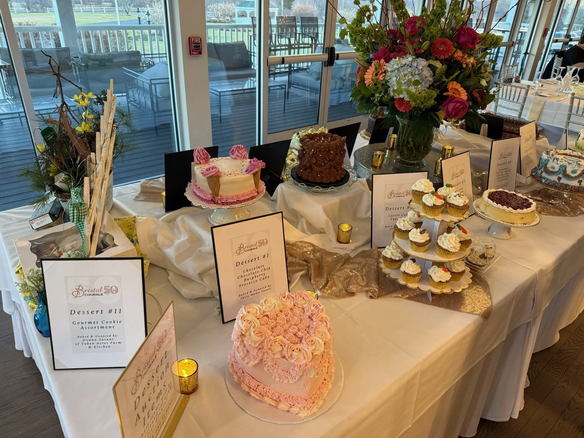 An indoor dessert display table with pink and white cakes, cupcakes, and dessert signs, decorated with a large colorful bouquet of flowers and small candle holders, with glass doors and seating area visible outside.