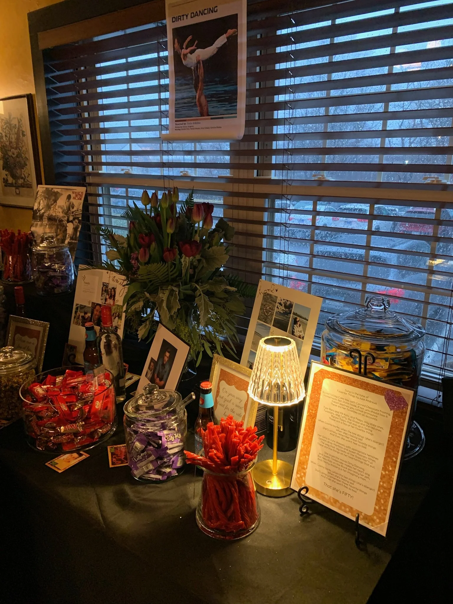 A table with various snacks, photographs, and informational signs illuminated by a small table lamp, with a large window with wooden blinds and a poster of a gymnast hanging on the wall in the background.