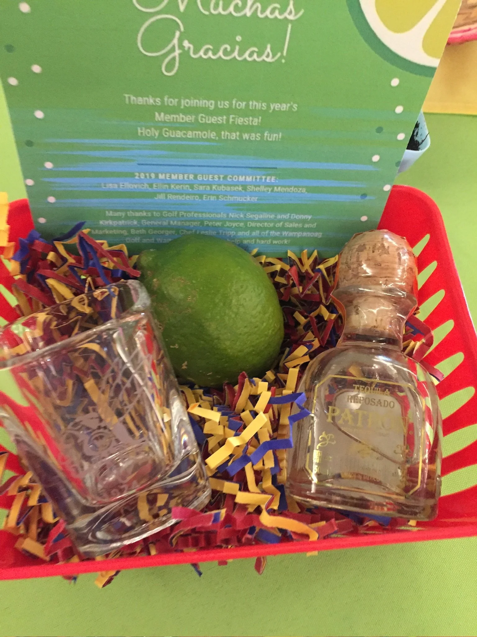 A thank you note for guests at a fiesta, with a lime, small bottles of tequila, and decorative paper shreds inside a red basket.