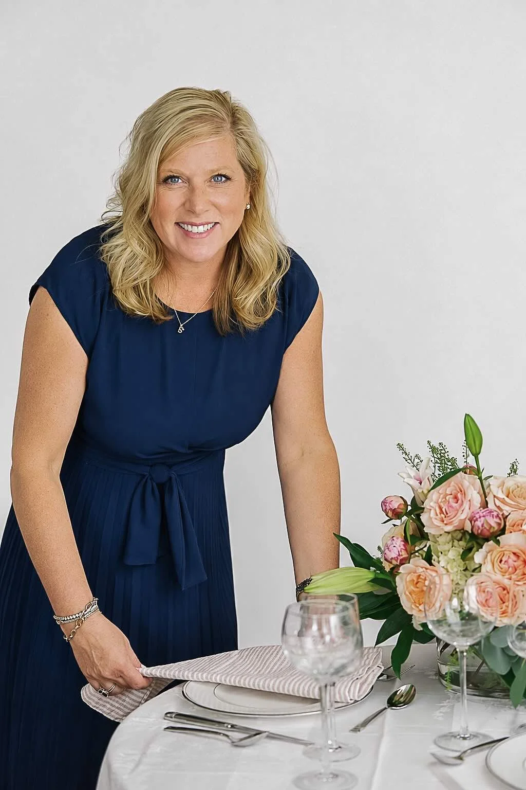 A woman with blonde hair, wearing a navy blue dress, smiling, standing by a table set with a floral centerpiece, glasses, and silverware.