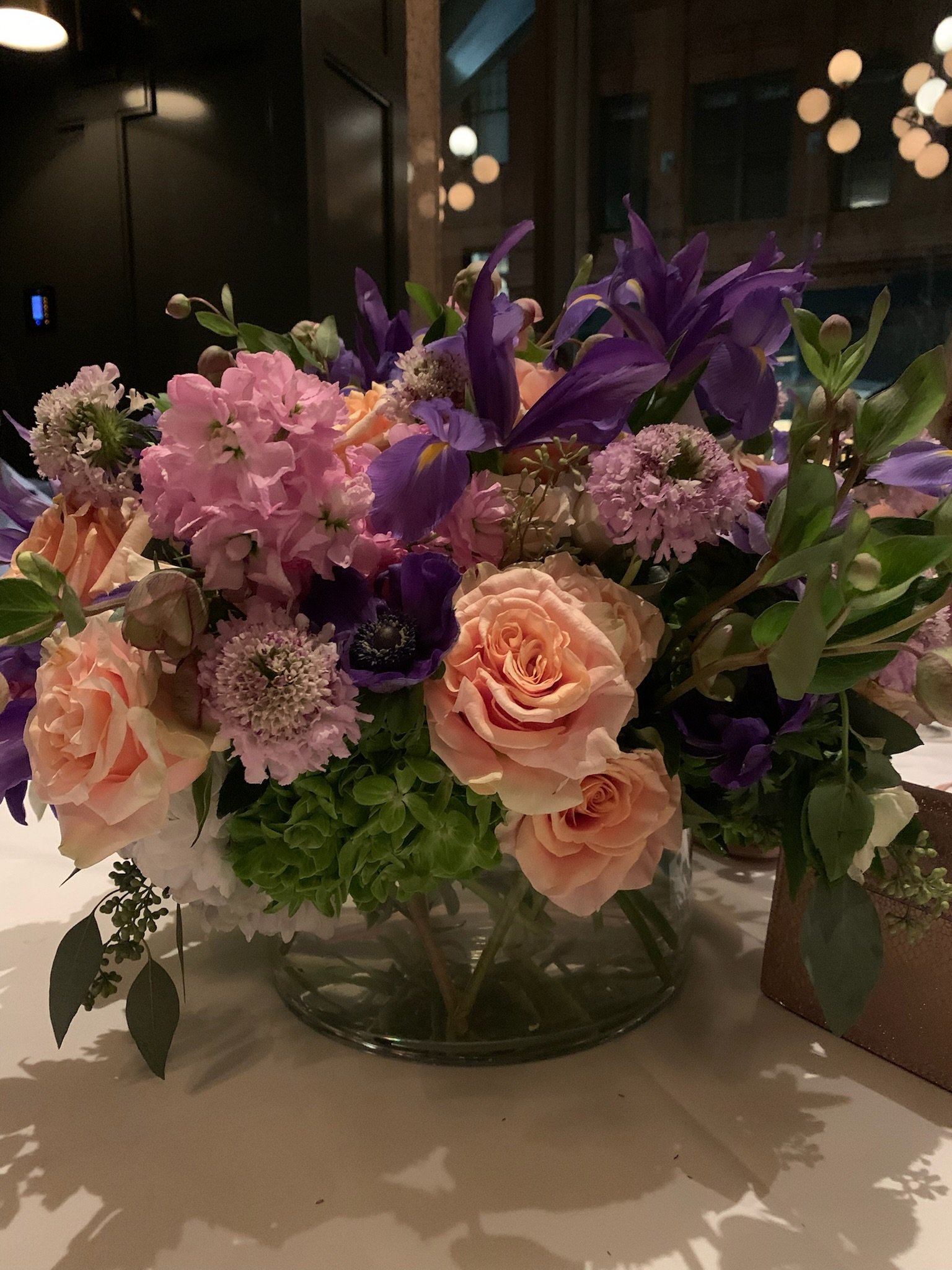 A large floral arrangement with purple, pink, and peach flowers, placed in a glass bowl on a table, with a dark background and round hanging lights.