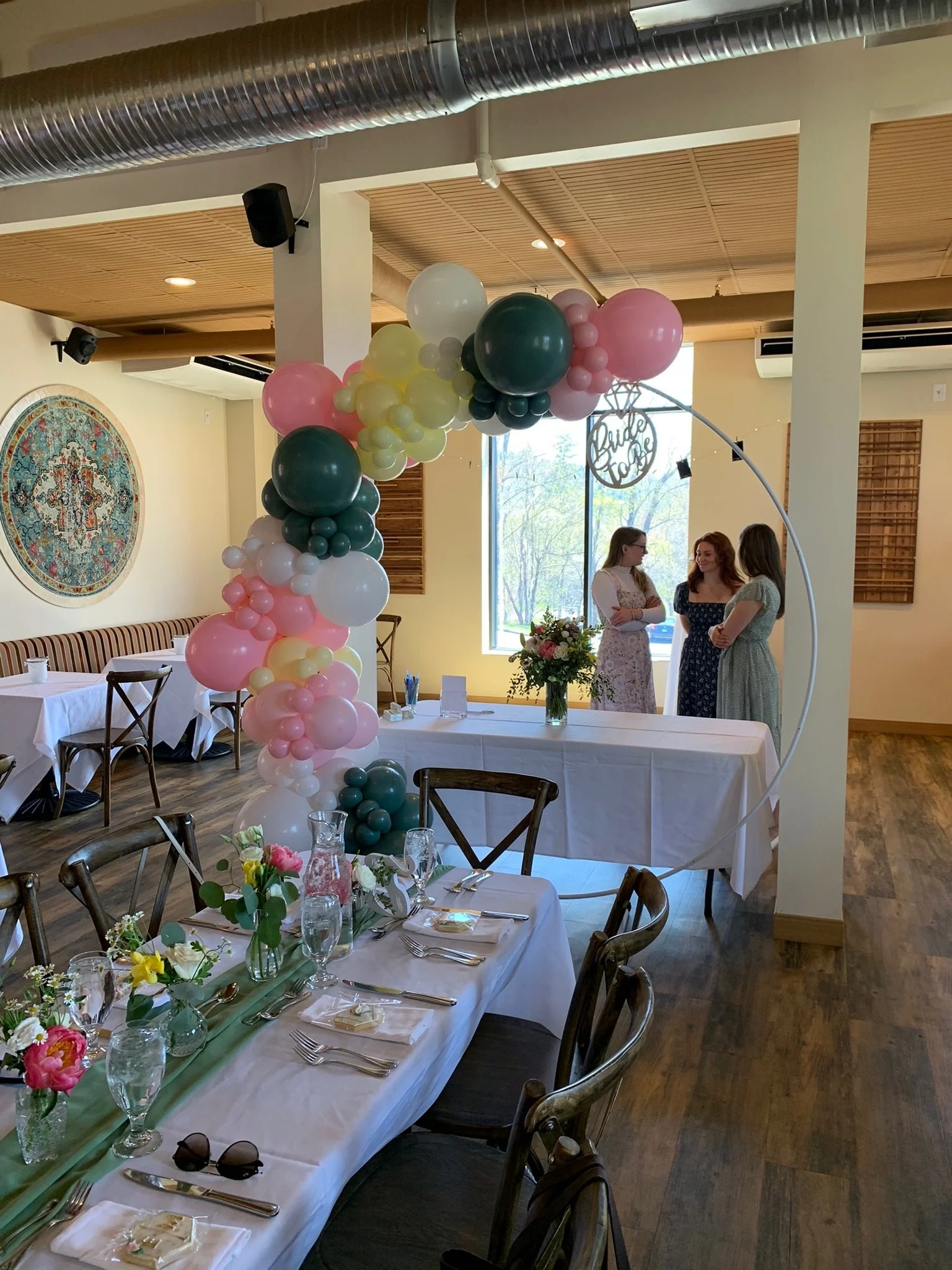 Indoor celebration venue decorated with a balloon arch in pink, white, yellow, and dark green. There are tables set with white tablecloths, floral centerpieces, glassware, and utensils. In the background, three women are standing near a window, engaged in conversation.