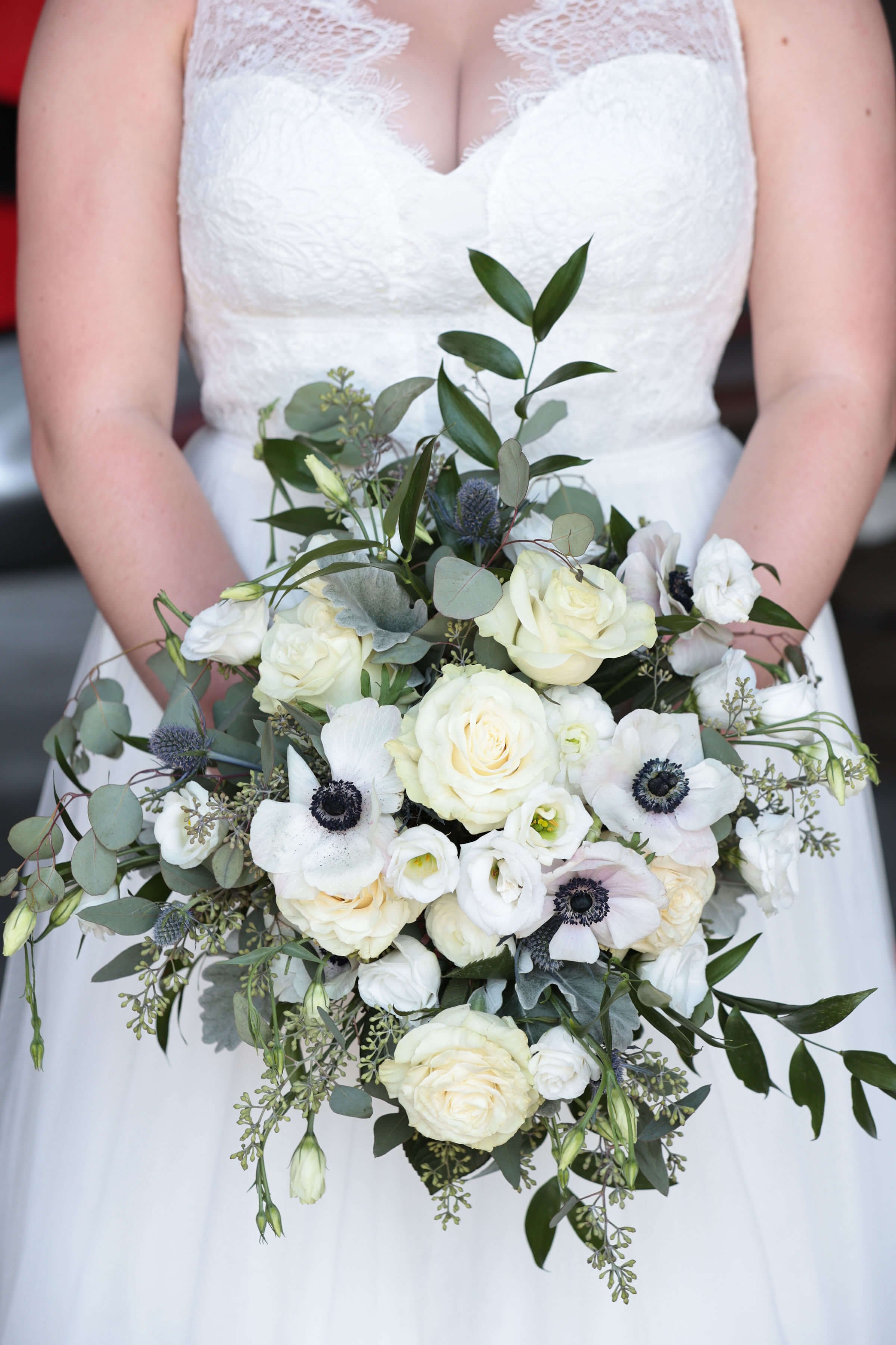 A woman in a white lace wedding dress holds a bouquet of white and green flowers, displayed in front of her chest.