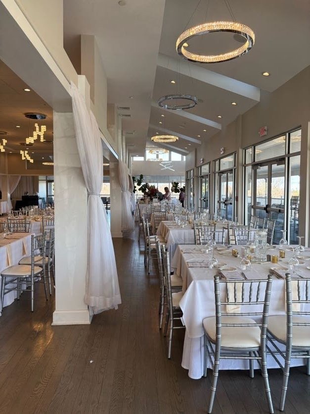 Elegant indoor dining area with white tablecloths, silver chairs, and large windows, set up for a formal event or wedding reception.