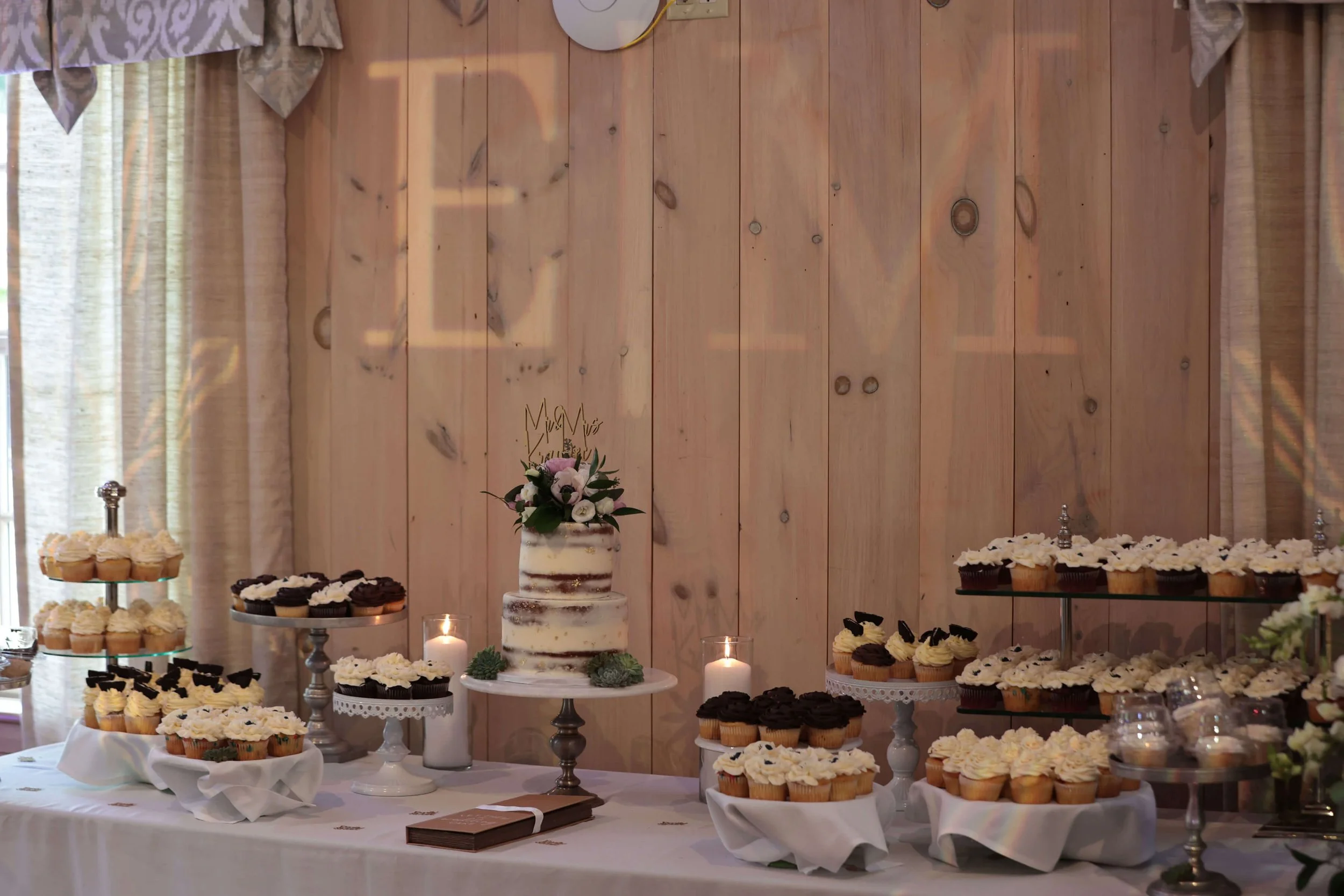 A dessert table at a celebration event with a white and brown rustic theme. The table has a layered wedding cake with flowers on top, numerous cupcakes with white and black frosting, and lit candles. The backdrop is a wooden wall with natural light coming through a window with curtains.