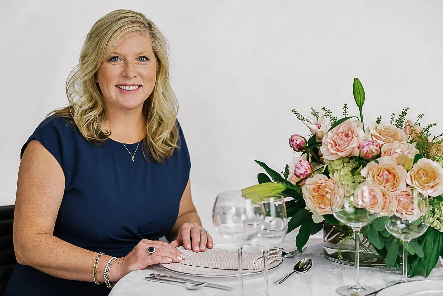 A woman with blonde hair, wearing a navy blue dress, sitting at a table decorated with a large bouquet of peach and pink flowers, clear glasses, and silverware.