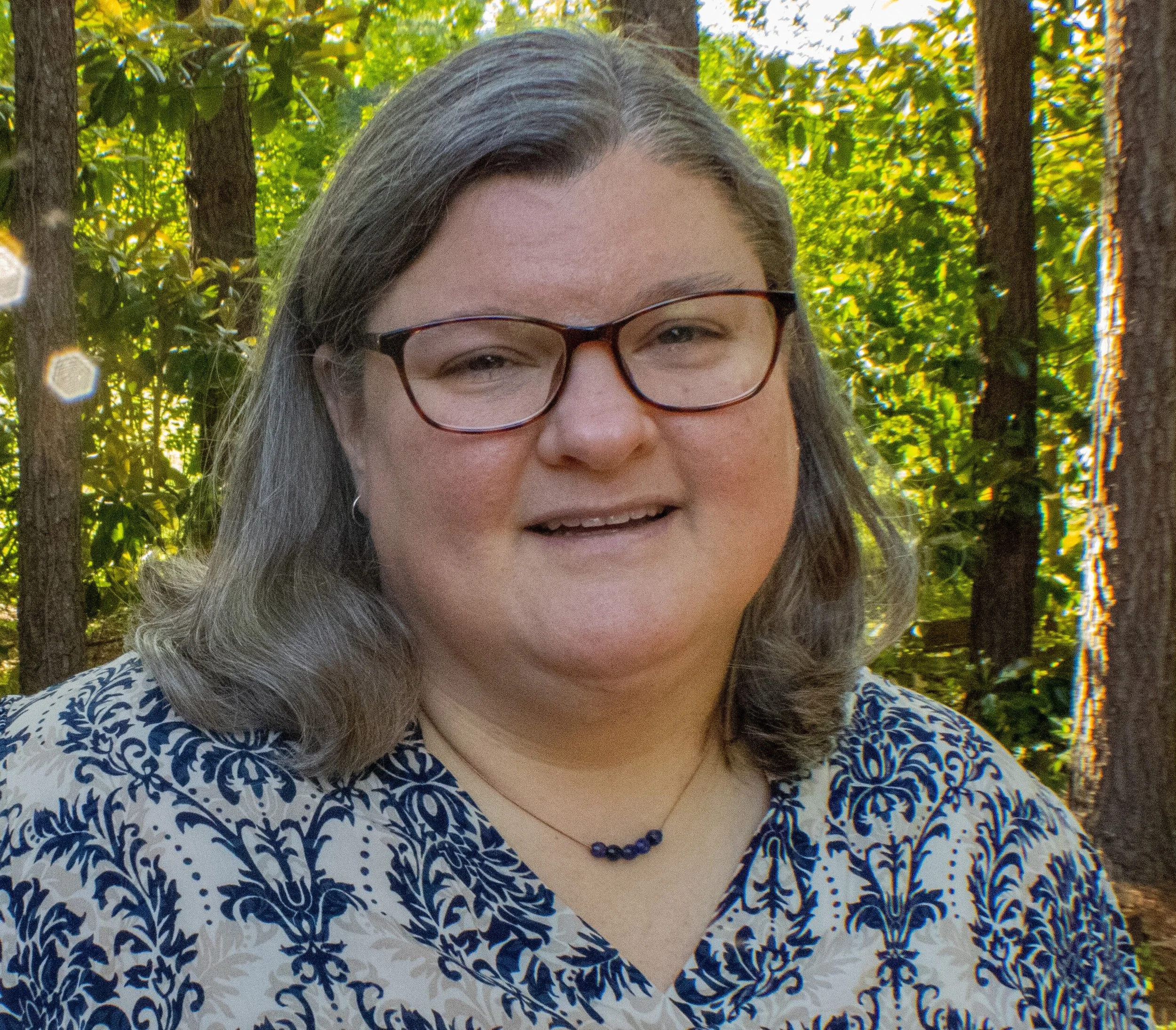 A woman with glasses and long hair smiling outside in a wooded area with green leaves and trees.