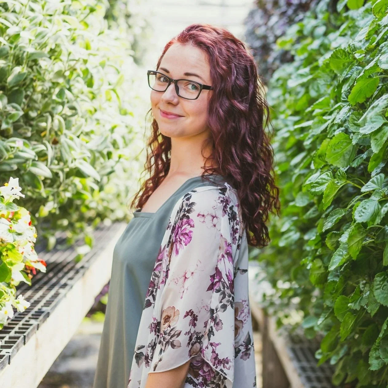 A woman with glasses and long curly hair smiling in a greenhouse surrounded by lush green plants and flowers.