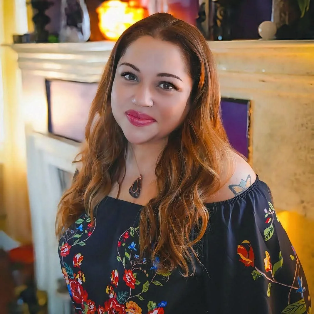 A woman with long wavy auburn hair smiling, wearing a dark floral off-shoulder dress, standing in a cozy room with warm lighting, a fireplace, and colorful decor.