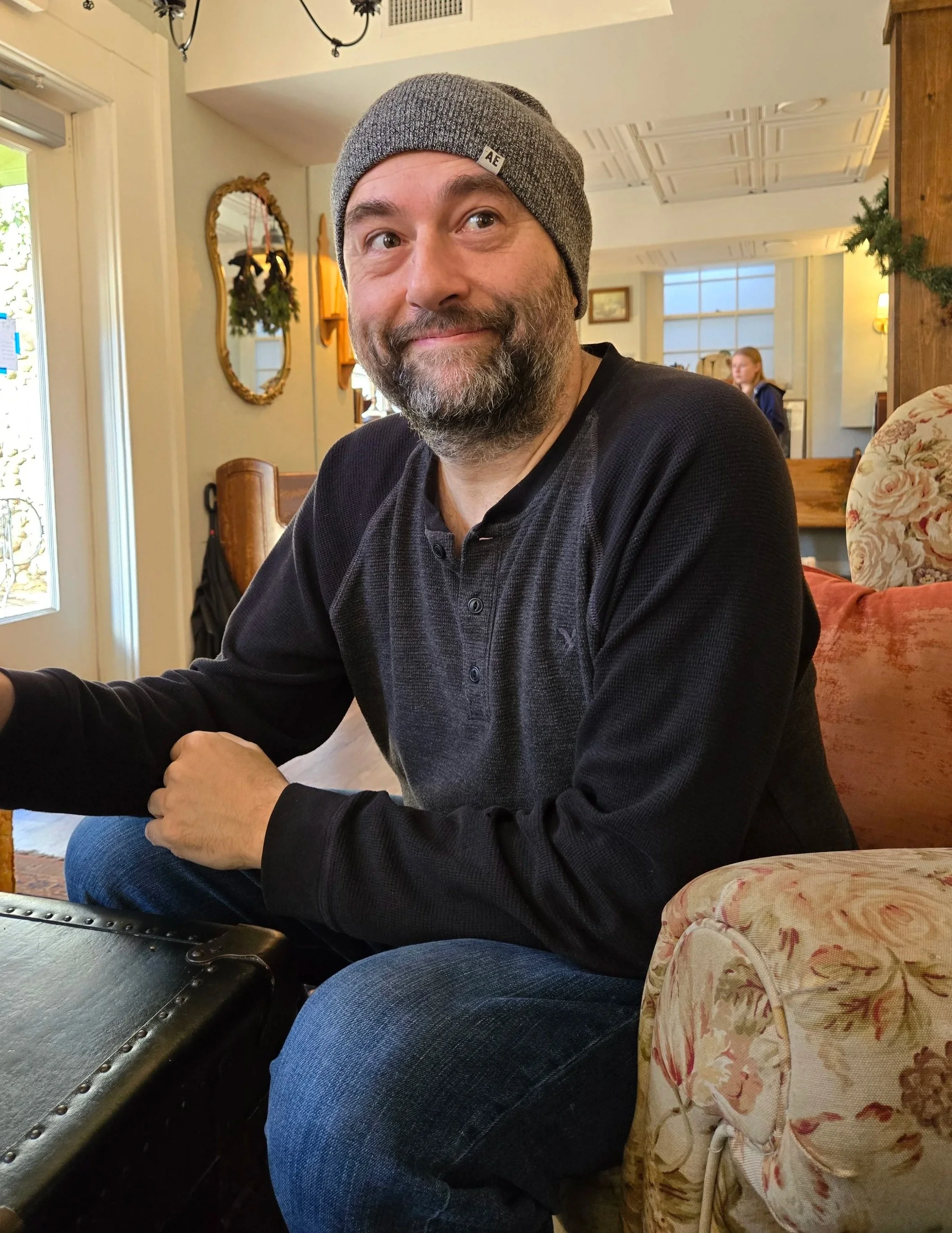 A man with a beard and wearing a gray beanie hat, smiling, sitting indoors at a table.