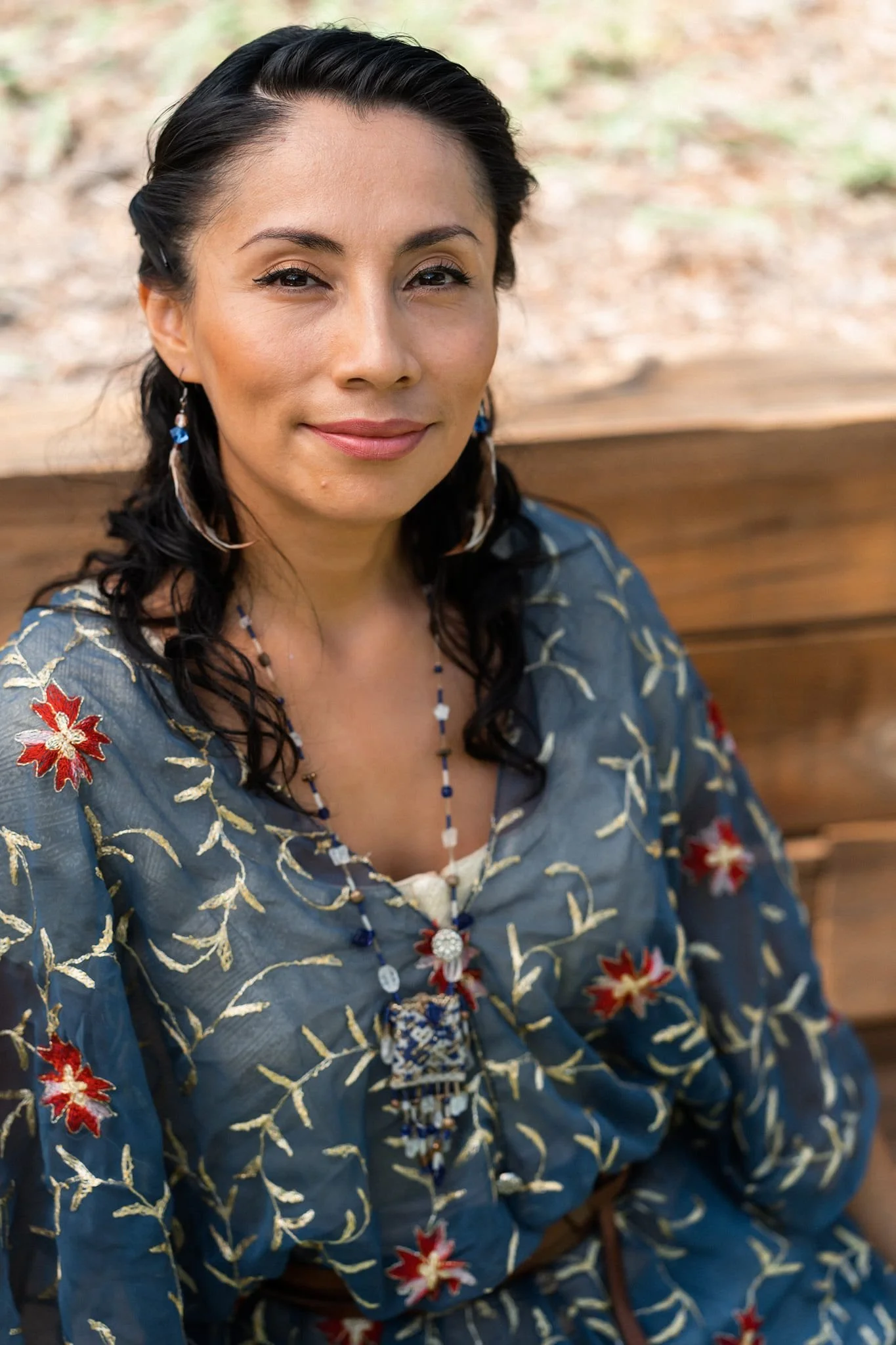 A woman with dark curly hair, wearing a blue blouse with red and white floral embroidery, and beaded jewelry, sitting outdoors on a wooden bench.