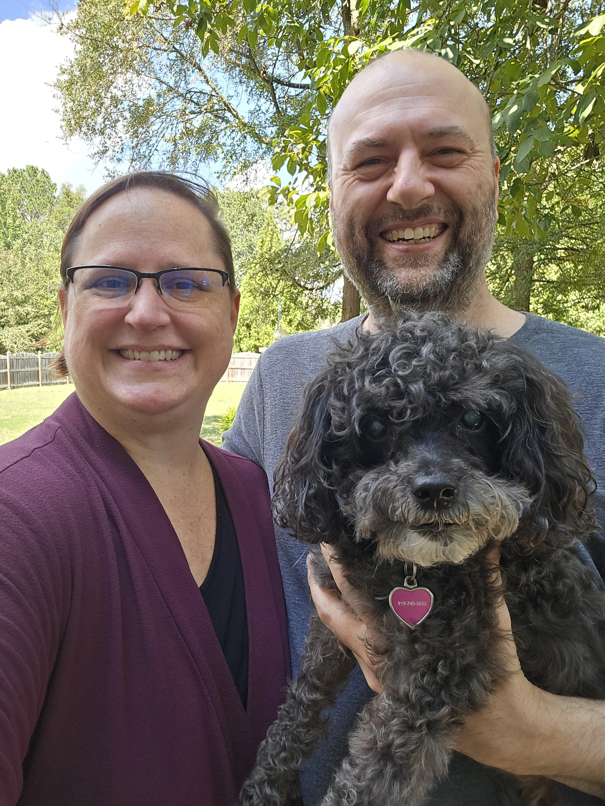 A smiling woman in glasses and a maroon blazer taking a selfie outdoors on a sunny day, with two smiling men and a curly-haired dog with a pink heart-shaped collar in the foreground. The background shows green trees and a blue sky.
