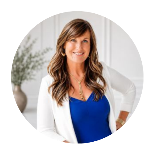 A woman with long brown hair wearing a blue top, white blazer, and jewelry, smiling at the camera inside a bright room with a plant in the background.