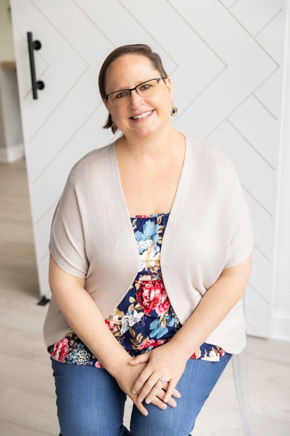 A woman with short brown hair, glasses, and earrings, smiling and sitting in front of a white wood-paneled wall.