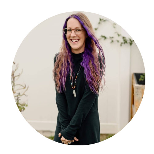 A woman with long, wavy hair dyed purple at the ends, wearing glasses, a black top, and a necklace, smiling and standing outdoors in front of a white wall and some plants.
