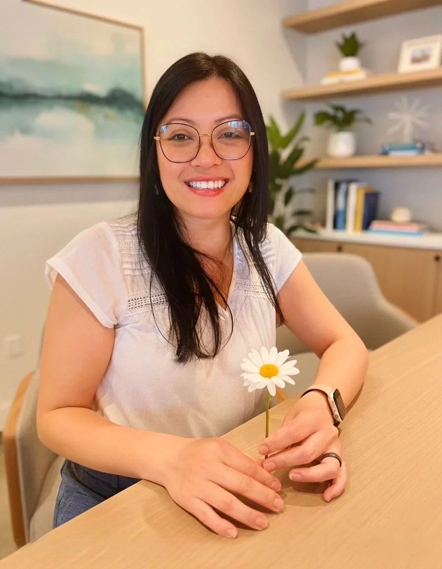 A woman with glasses and a white blouse smiling at the camera while holding a daisy flower in her hand, seated at a wooden table in a cozy room with shelves and a painting in the background.