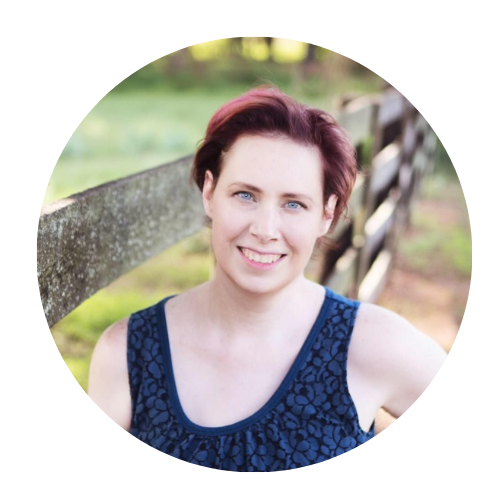 A woman with short reddish-brown hair, blue eyes, and fair skin, smiling outdoors near a wooden fence and greenery in the background.