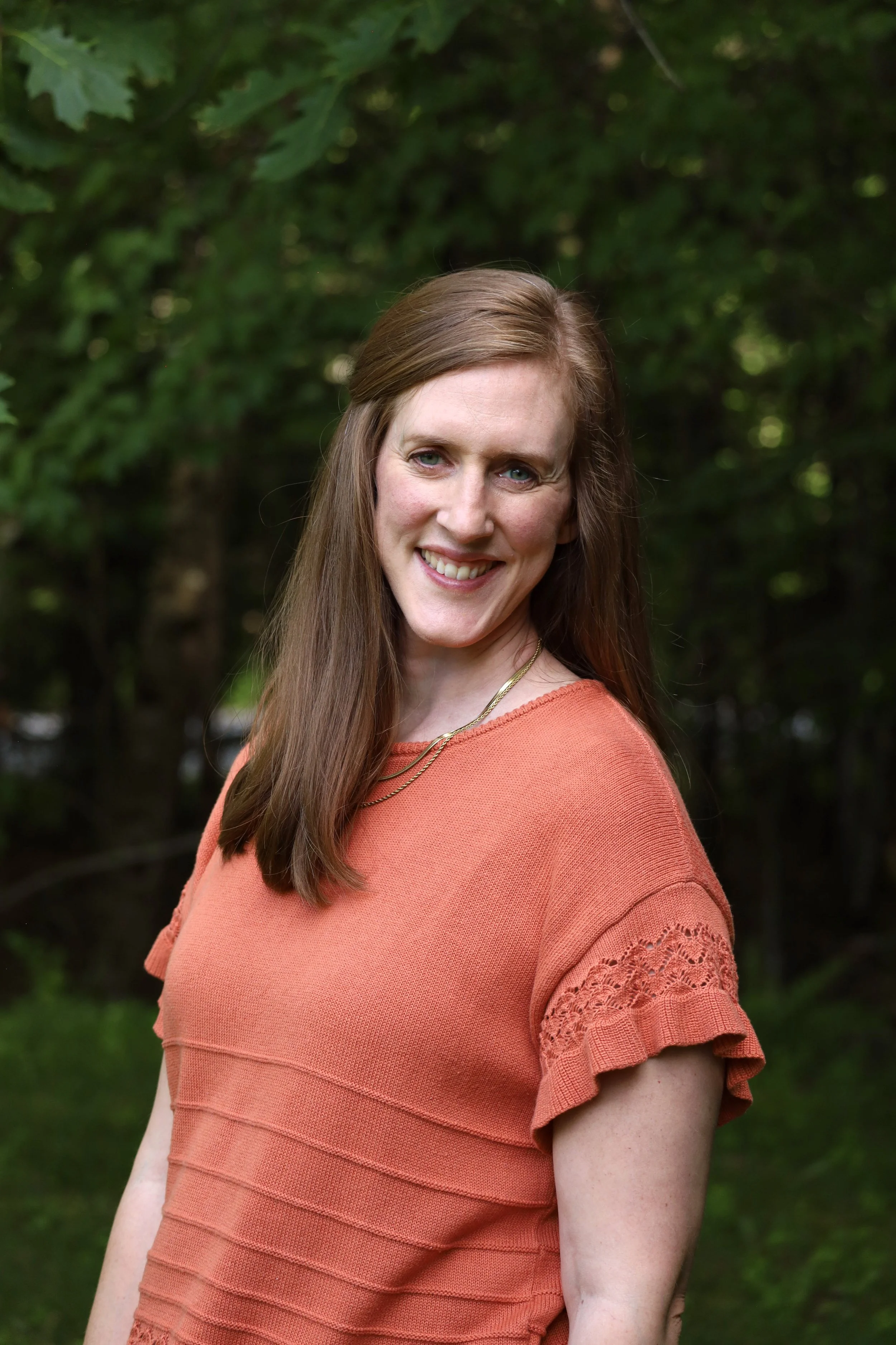 A woman with long brown hair and blue eyes smiling outdoors with green trees in the background, wearing an orange styled top with lace details on the sleeves and a gold necklace.