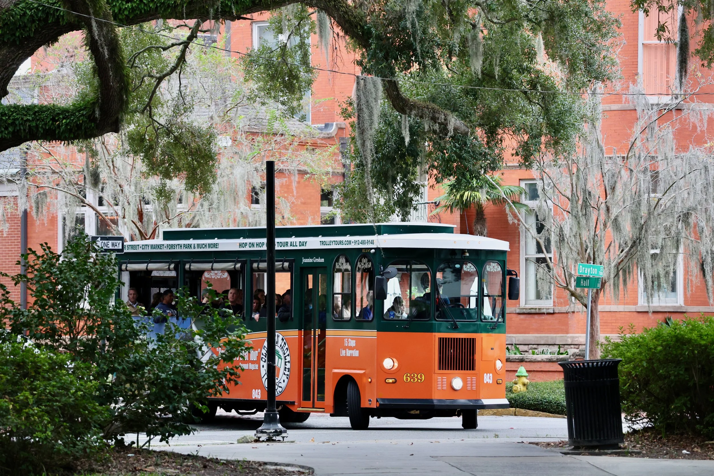 An orange trolley bus with passengers on board, traveling through a tree-lined urban area on Drayton Street in Atlanta, Georgia. There are green trees with Spanish moss hanging from the branches, brick buildings in the background, street signs, and a