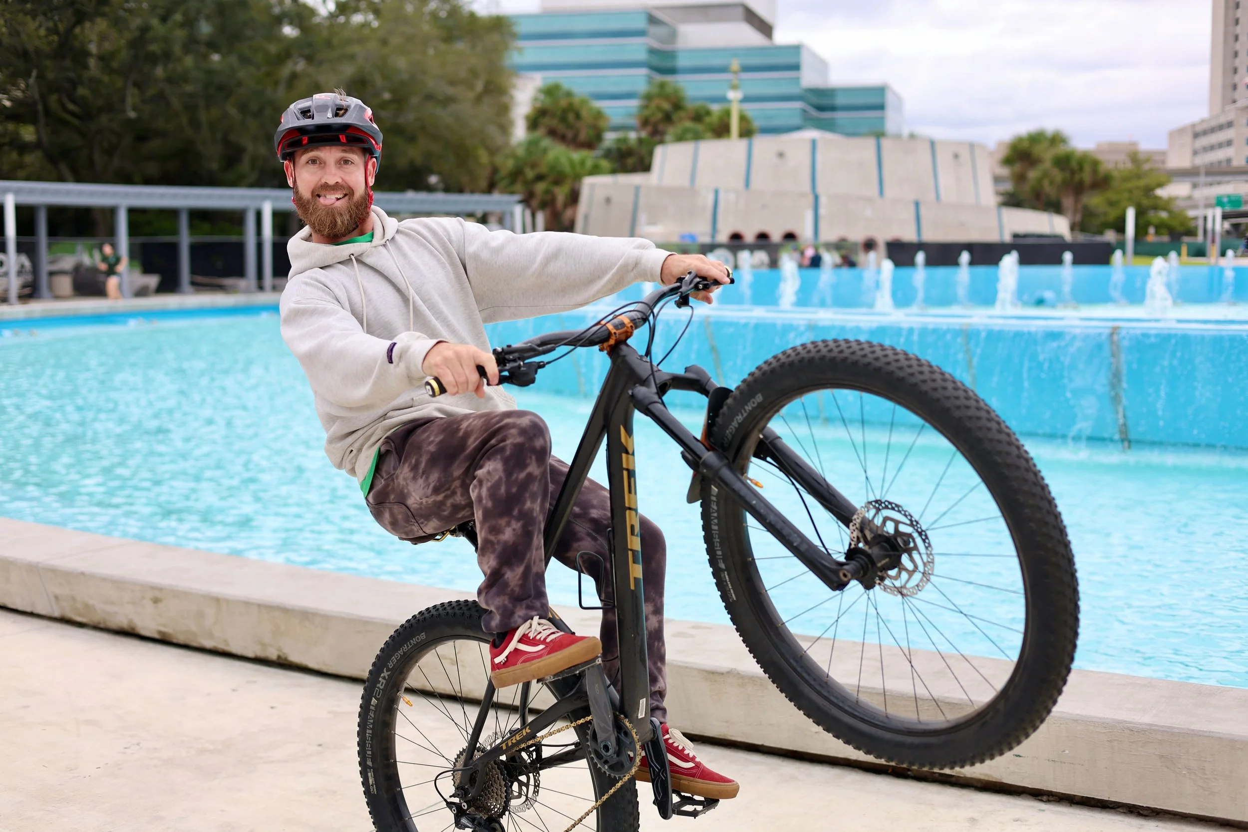 A man with a beard wearing a bike helmet, hoodie, plaid pants, and red sneakers riding a mountain bike near a large blue water fountain at an outdoor urban park.