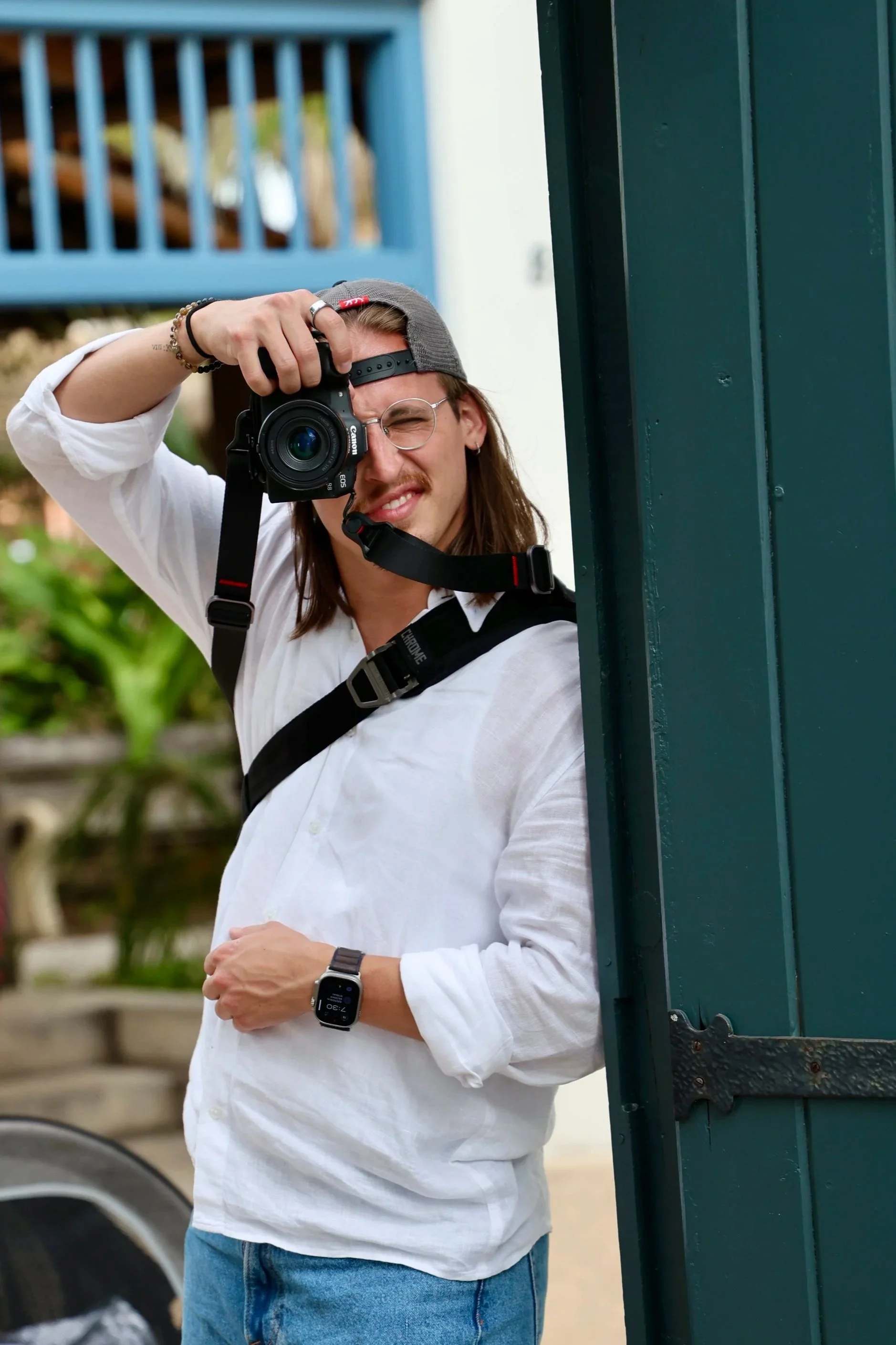 A man with long hair, glasses, and a gray cap is taking a photo with a Canon camera. He is wearing a white shirt and blue jeans, and has a smartwatch on his left wrist. He is standing outdoors near green-painted metal structures.