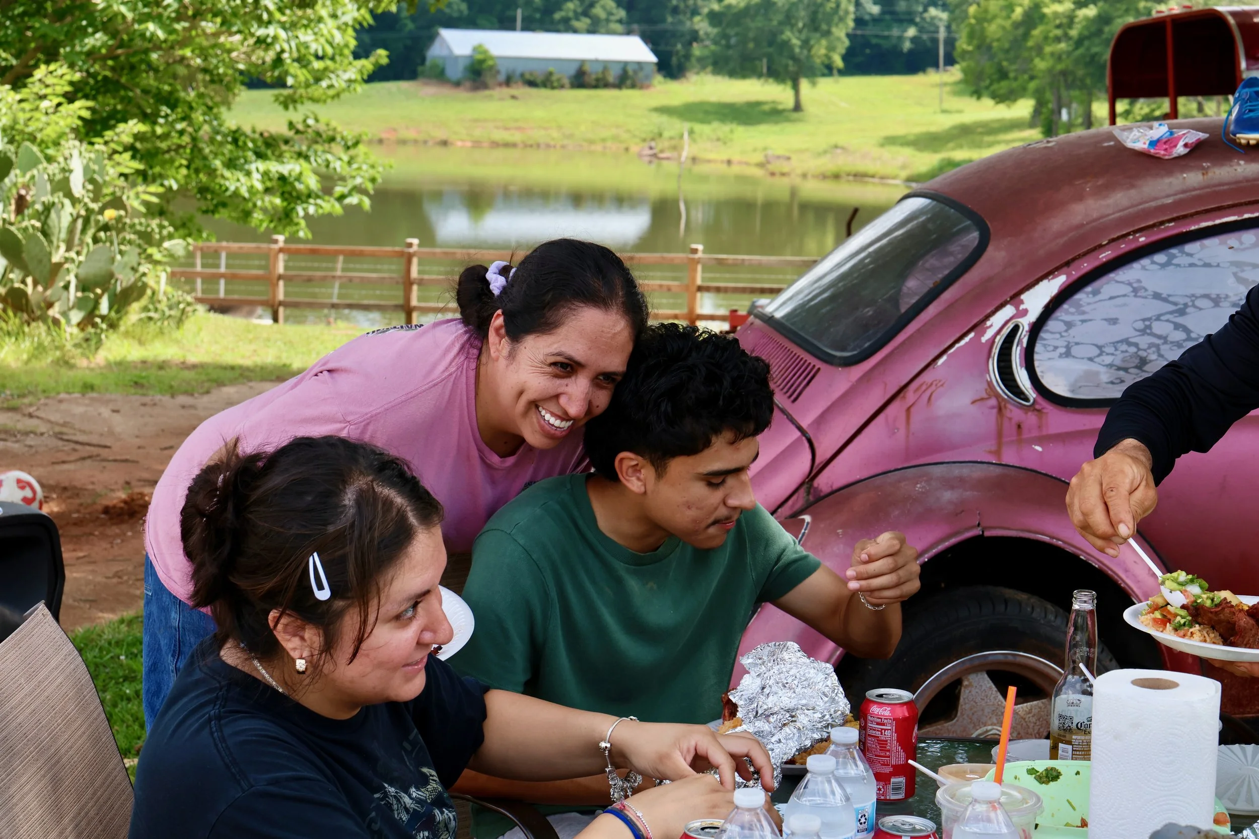 People gathered outdoors at a picnic table, with a body of water and green trees in the background. An old pink car is visible behind them.