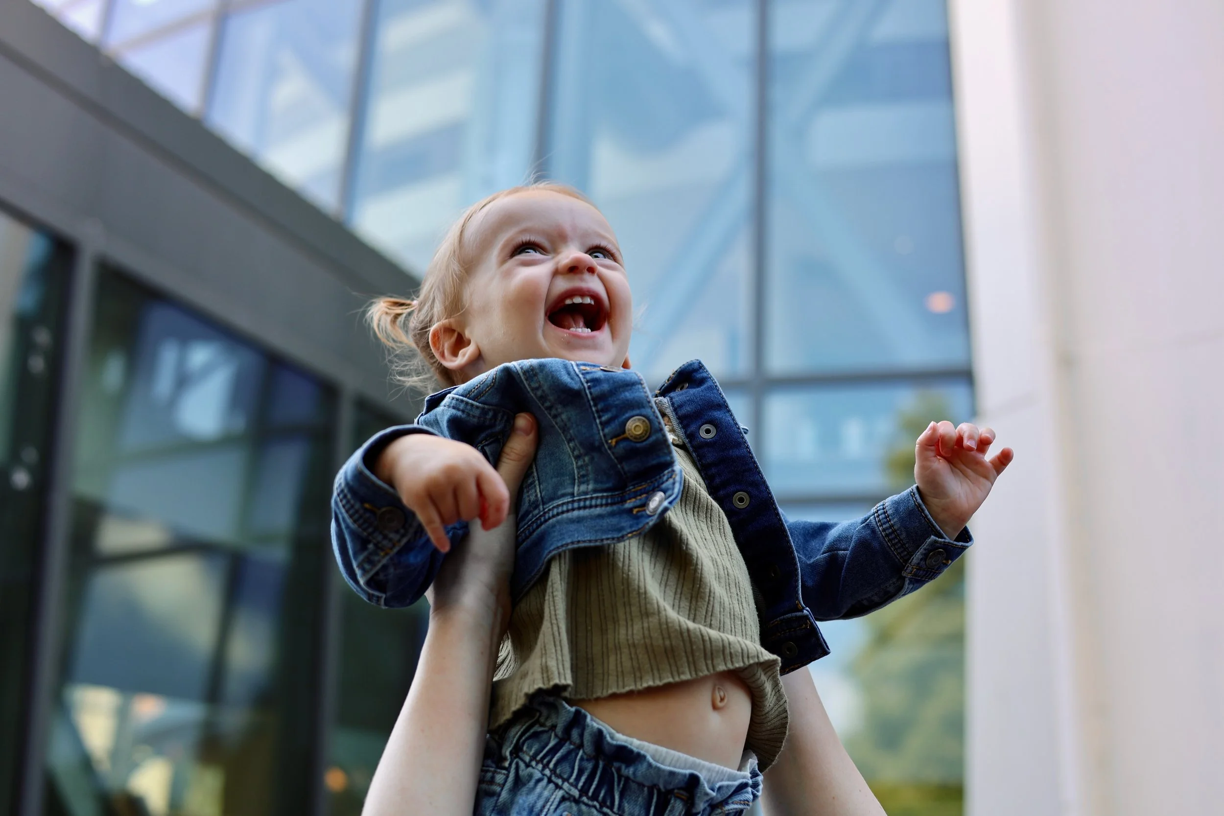A young child with blonde hair and a denim jacket being held up outdoors in front of a modern glass building, looking upwards with an excited expression.