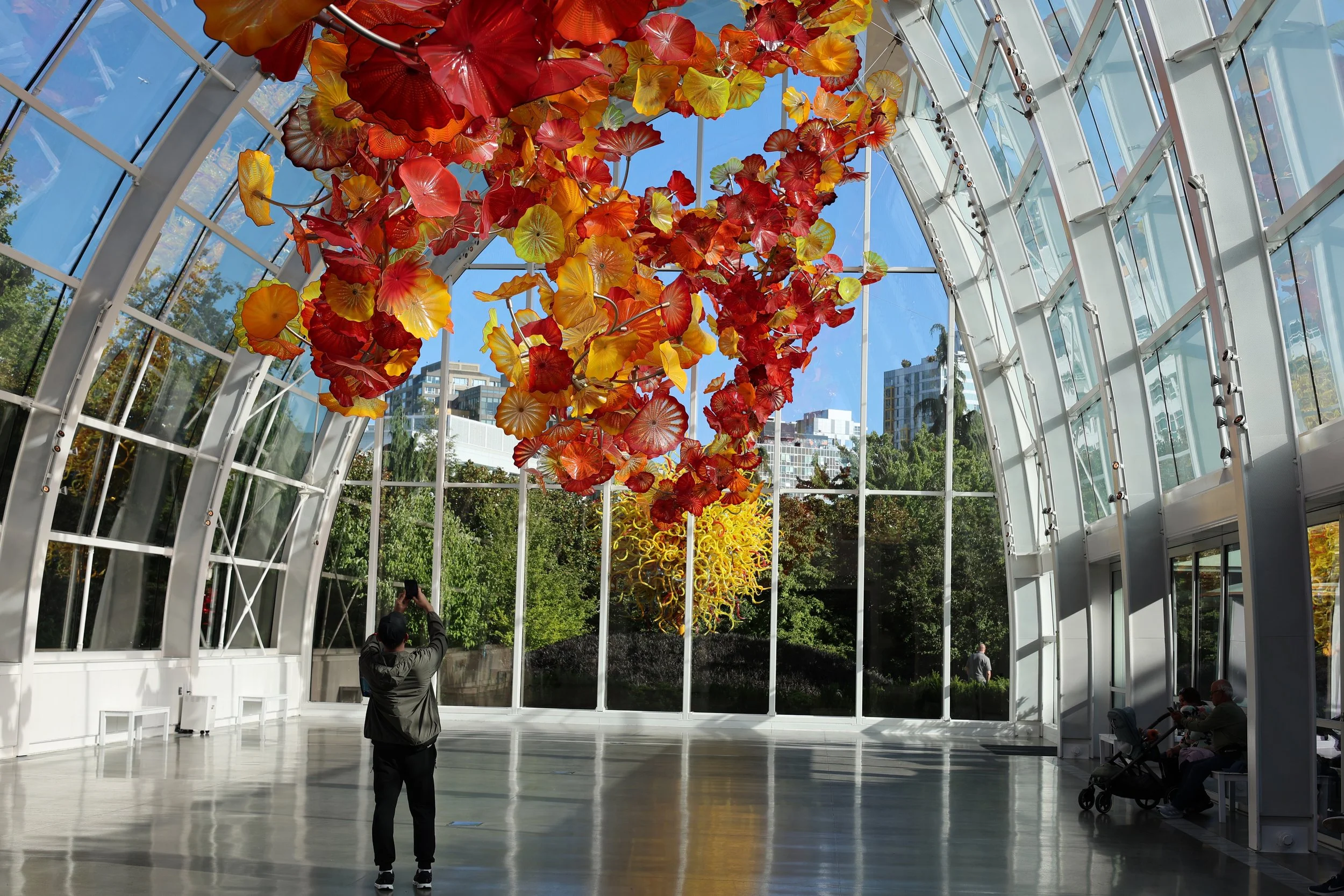 An interior view of a glass conservatory with a colorful hanging glass sculpture resembling flowers or leaves, featuring shades of red, orange, yellow, and green. Several people are visible inside, some standing, some sitting.