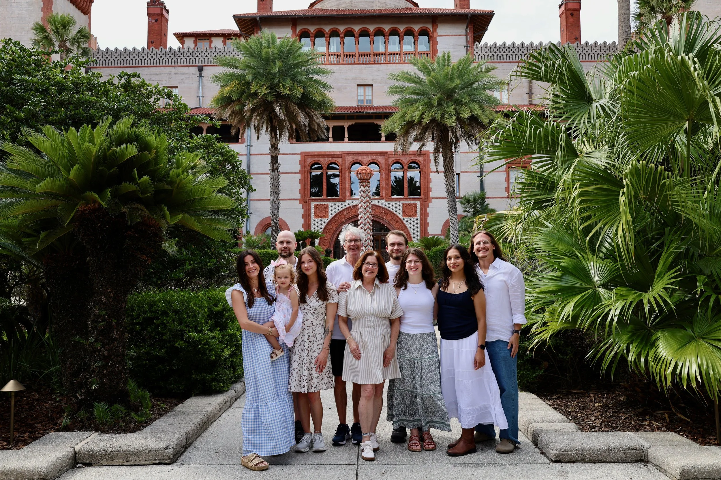 A group of people, including men, women, and children, standing together outdoors in front of a historic building with ornate architecture and surrounded by lush greenery and palm trees.