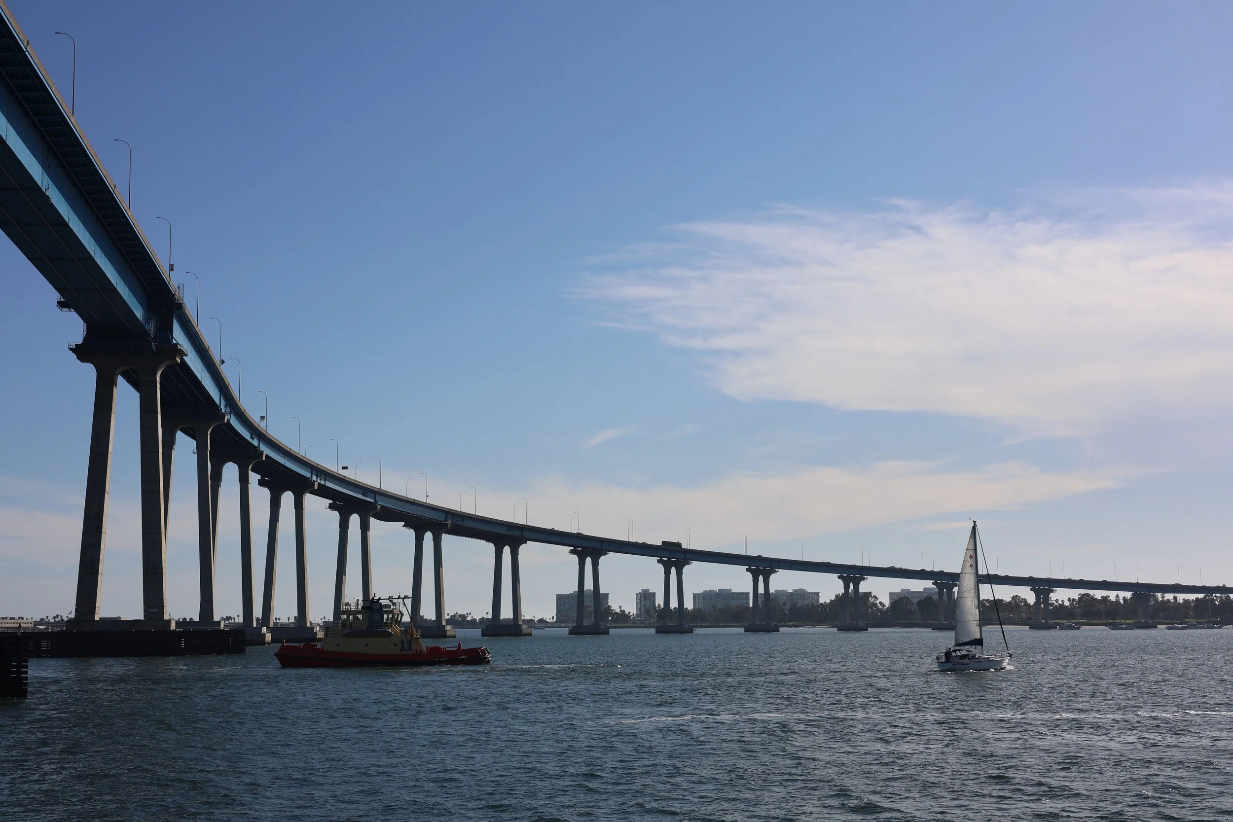 A bridge over water with sailboats and a tugboat in the foreground and buildings in the background under a partly cloudy sky.