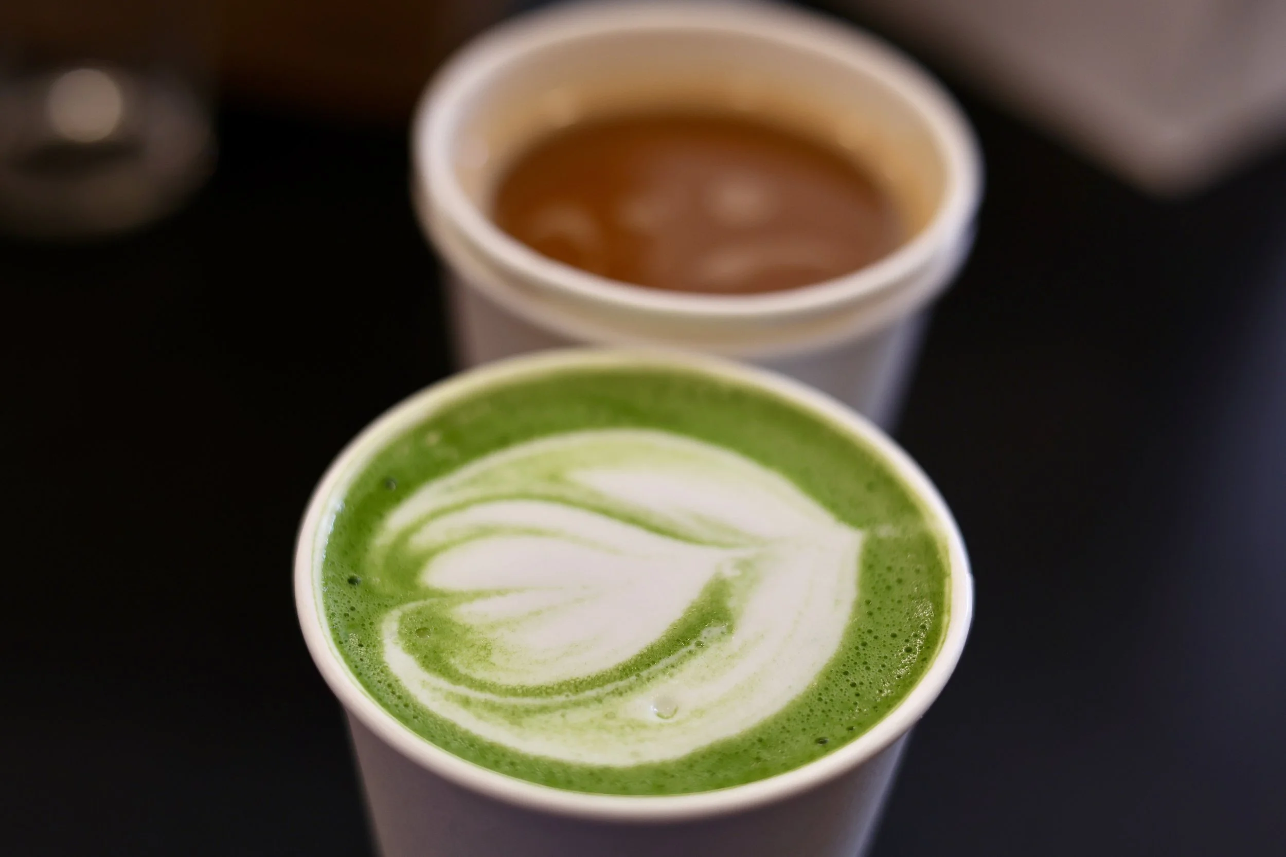 Close-up of a white cup with green matcha latte topped with white foam in a swirled design, with a blurred cup of coffee behind it.