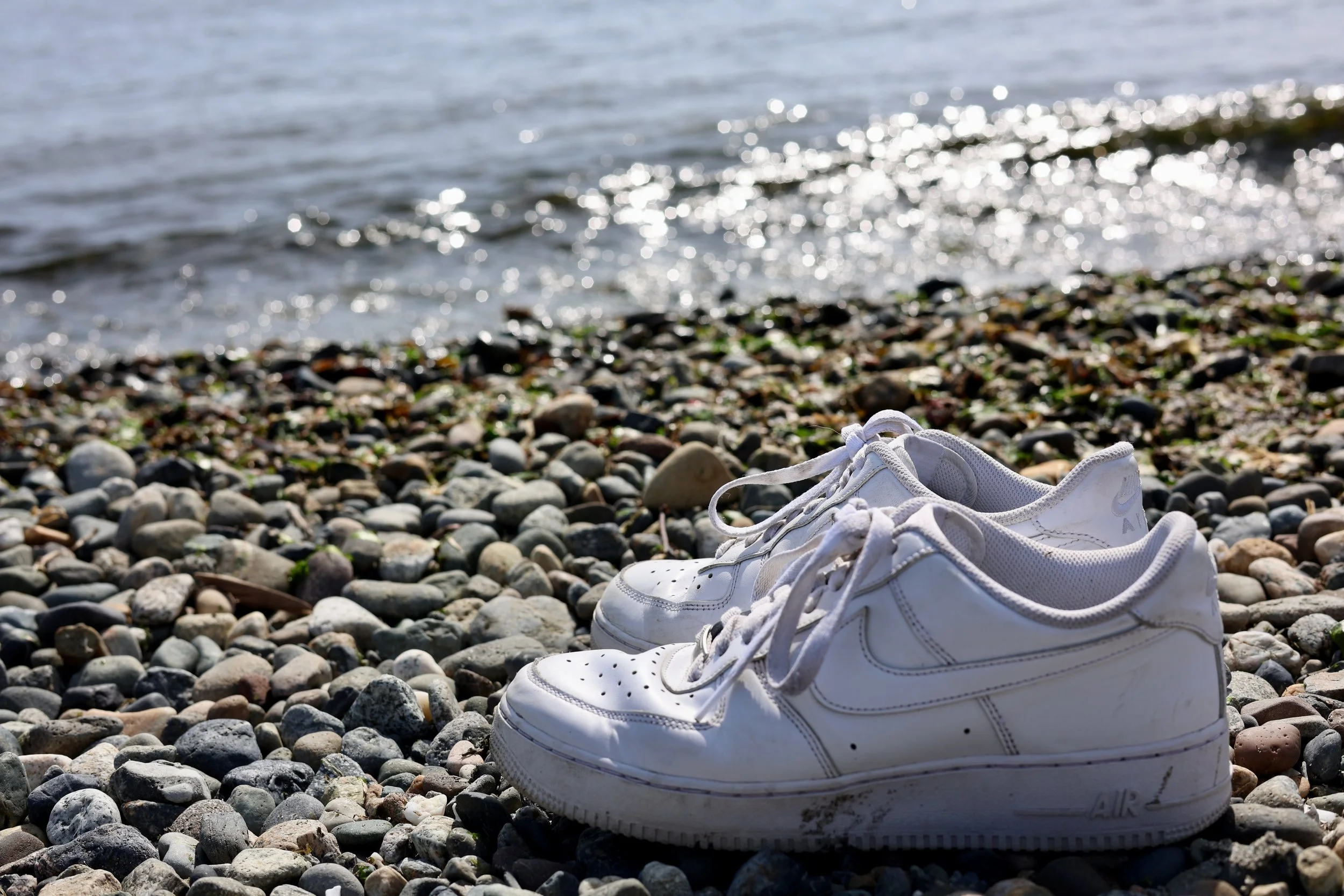 A pair of white athletic shoes on a rocky beach with water and sunlight in the background.