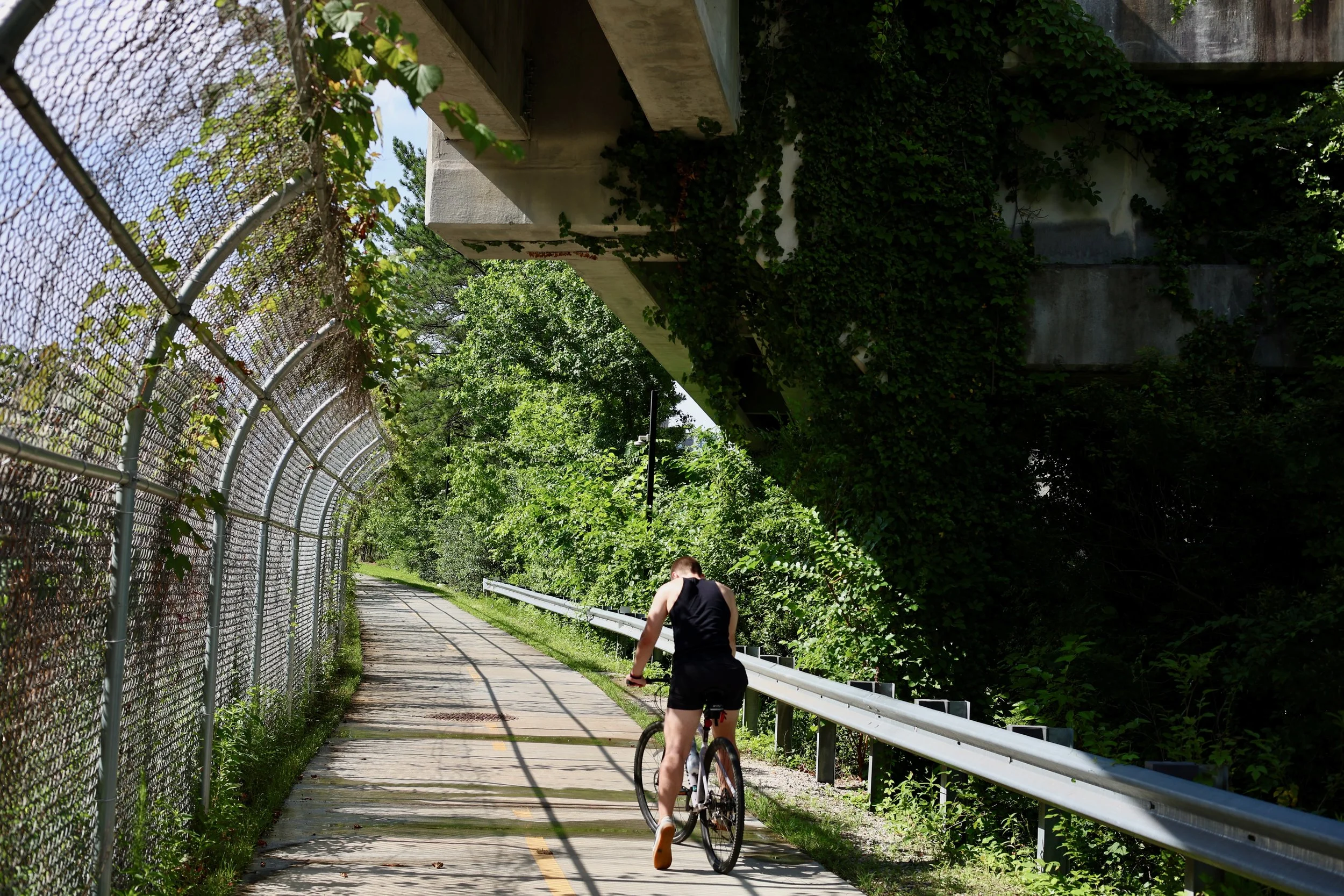 A person riding a bicycle on a paved trail alongside a chain-link fence covered with green plants. The trail is surrounded by trees and greenery, with a concrete structure and metal guardrail on the right side. The person is wearing a black tank top 