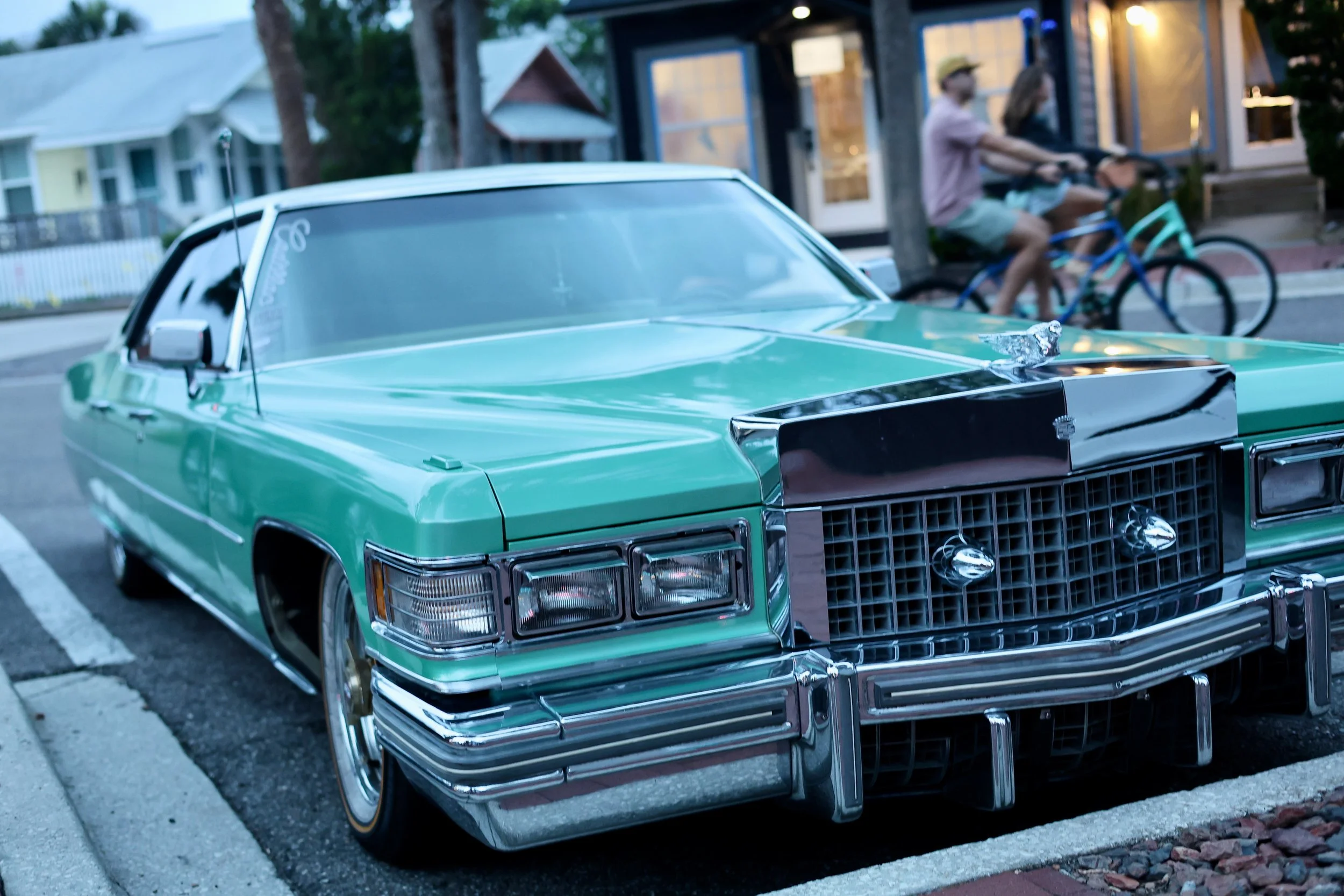 A vintage green Cadillac parked on the street with a house and people riding bicycles in the background during dusk.