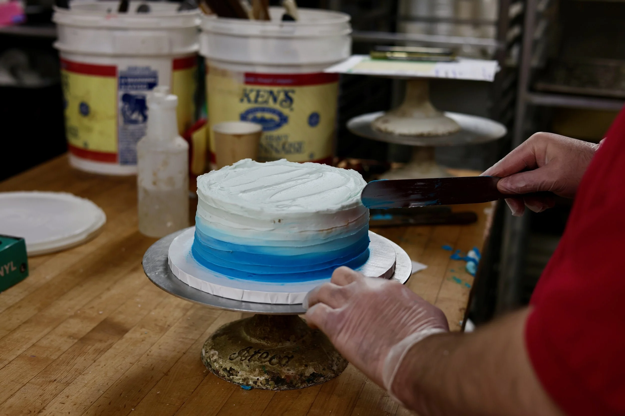 A person wearing gloves is applying white and blue icing to a cake on a wooden work surface, with bakery supplies and containers in the background.