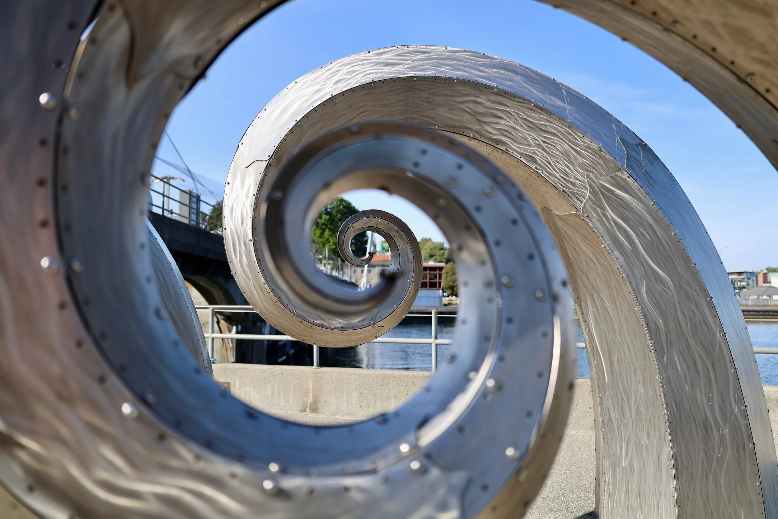 Close-up of a large metallic spiral sculpture outdoors, viewed from the side with buildings and water in the background.