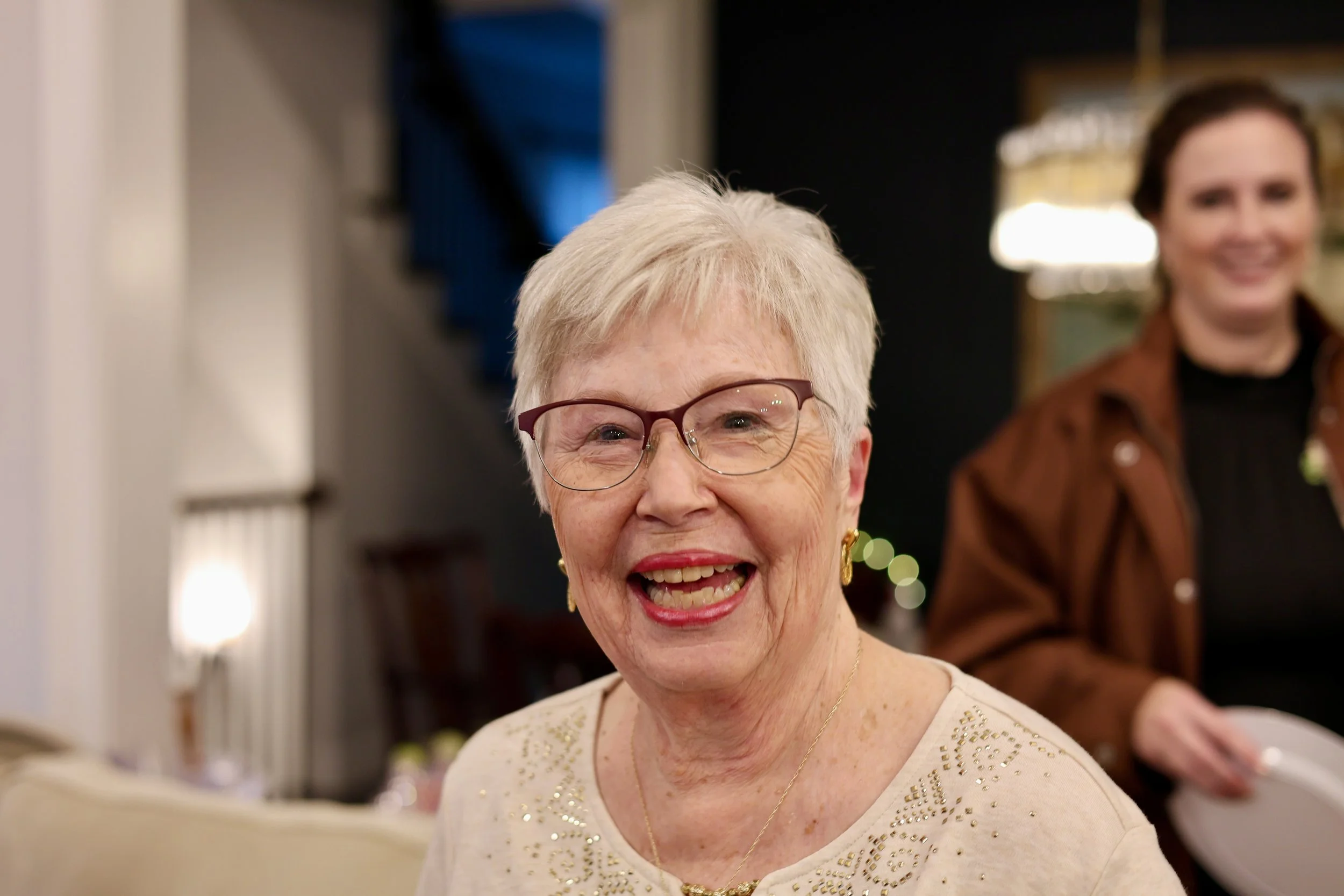 An elderly woman with short gray hair, glasses, and a beaming smile, wearing a cream-colored top with gold embellishments, at a social gathering.
