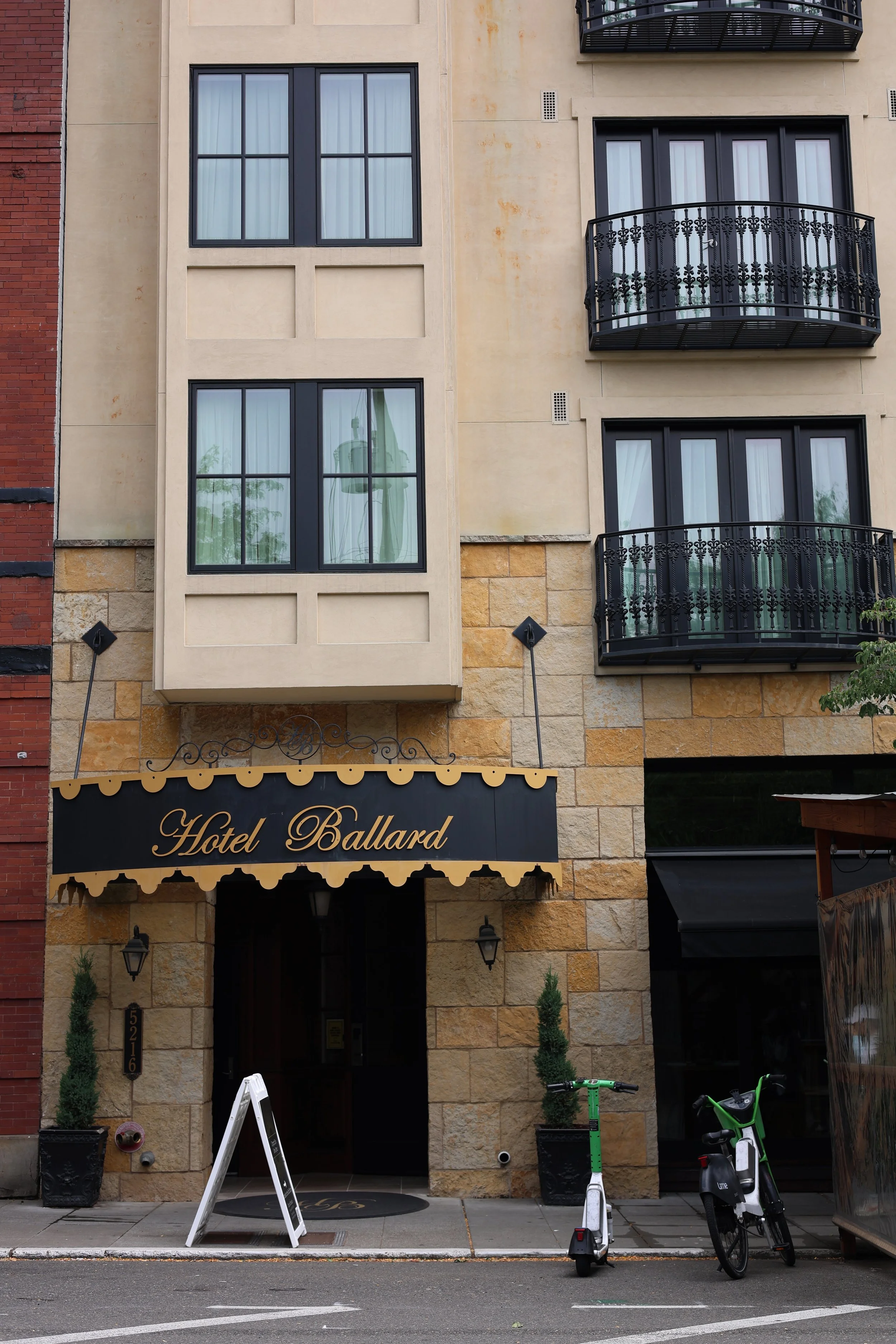 The front entrance of Hotel Ballard, with a black and gold sign above the doorway, and a black awning. The building has large windows with curtains, small black balconies, and a mix of beige and brick exterior. There are green bicycles and scooters parked outside, with a small whiteboard sign and potted plants near the entrance.