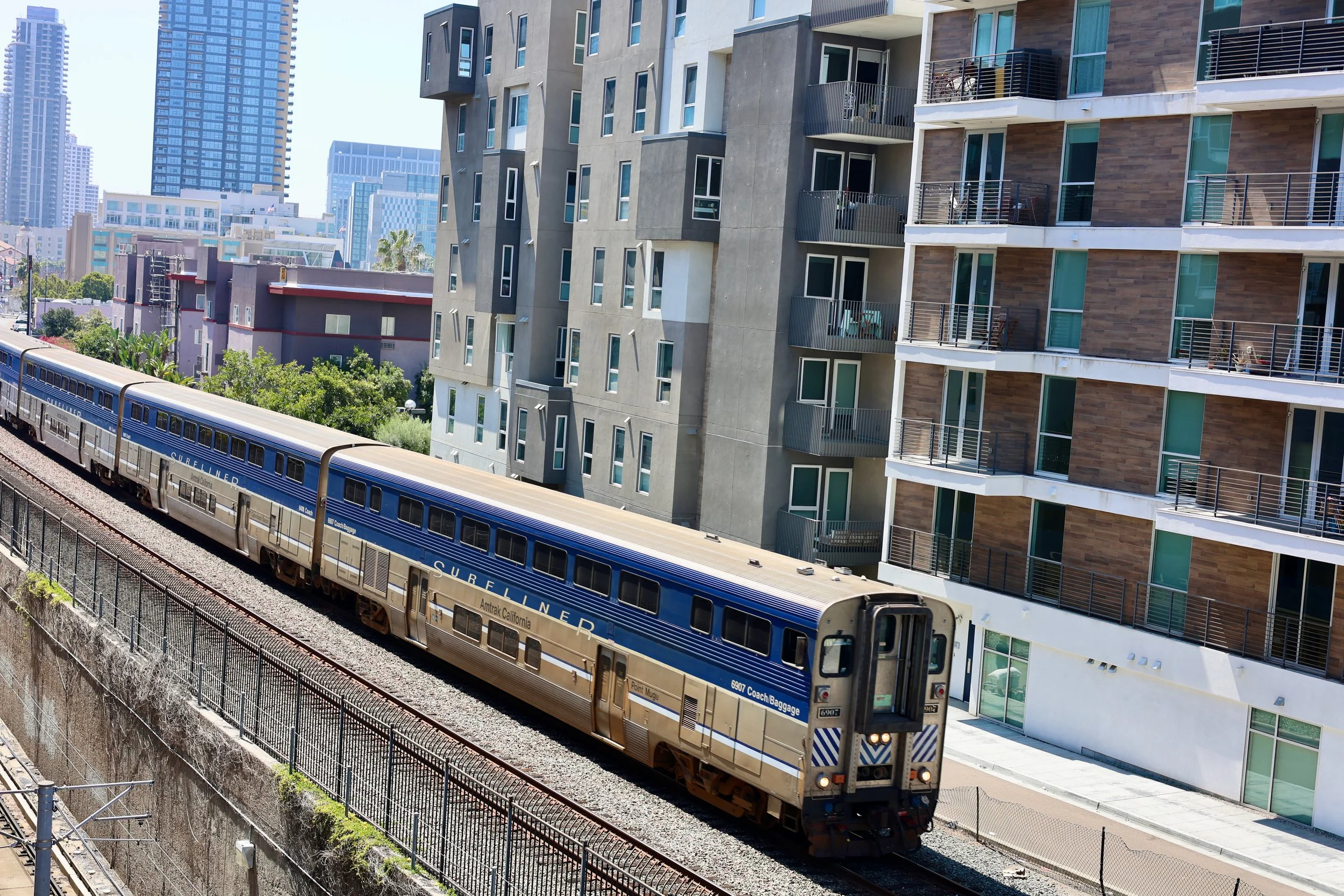 A passenger train with blue and silver colors moving on railway tracks in front of modern multistory apartment buildings in an urban area on a sunny day.