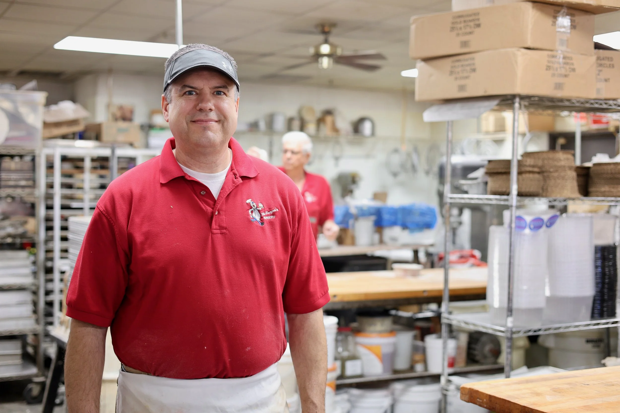 A man wearing a gray visor and a red polo shirt with a bakery logo stands in a bakery supply room. Behind him, shelves hold various boxes and bakery supplies, and other employees are visible in the background.