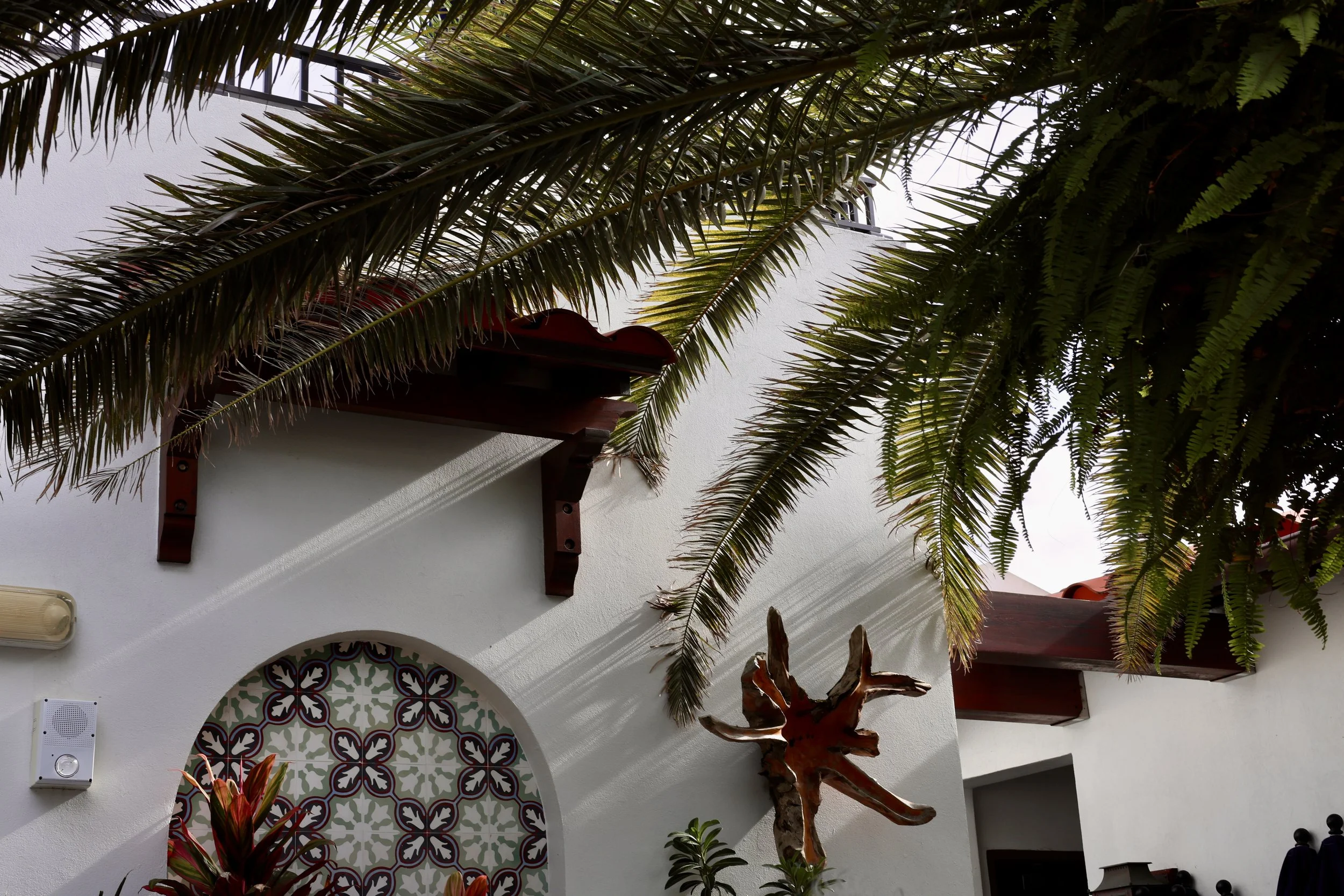 A white building with a decorative arch window featuring an intricate multicolored tile pattern. Tall palm trees and lush green plants surround the building. Part of a red-tiled roof and some wooden structures are visible.