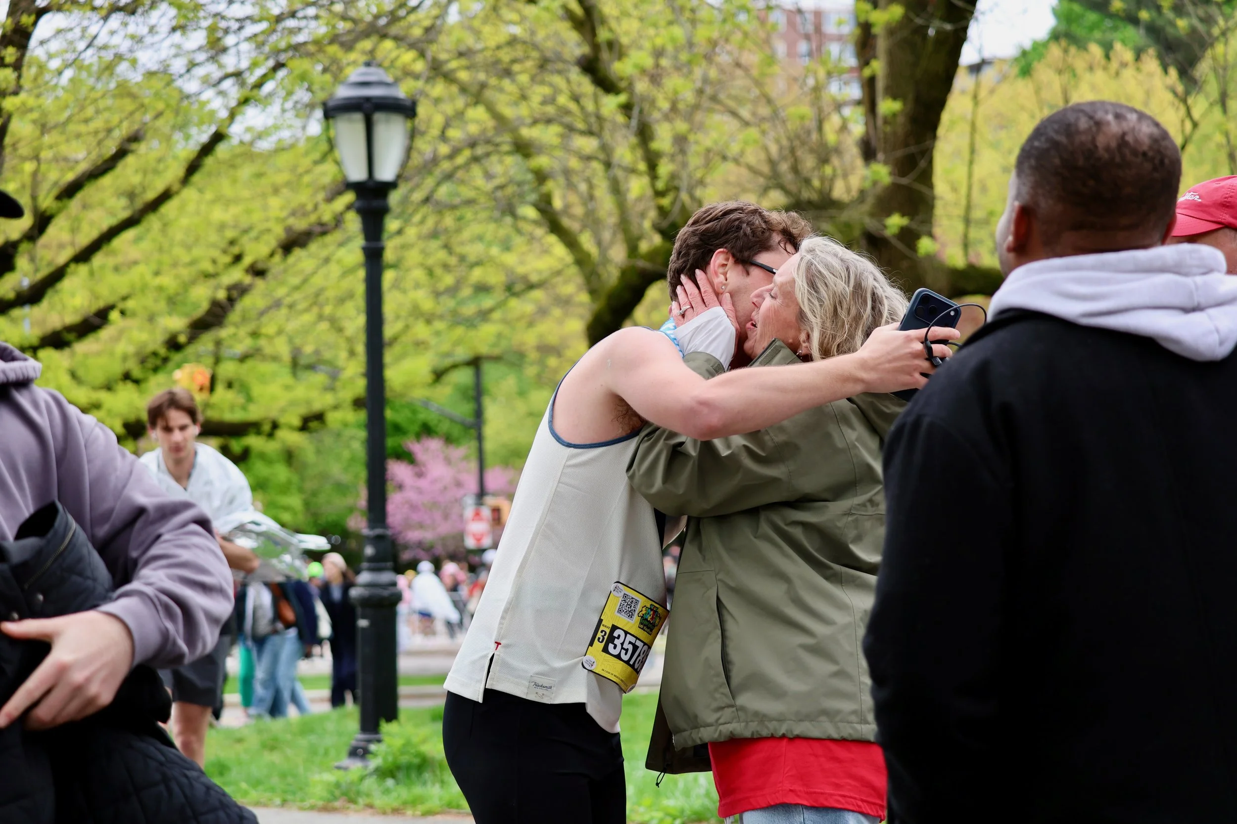 A mother and son embrace in Prospect Park after the Brooklyn Marathon, with participants and onlookers around, trees with green leaves, and a lamp post in the background.