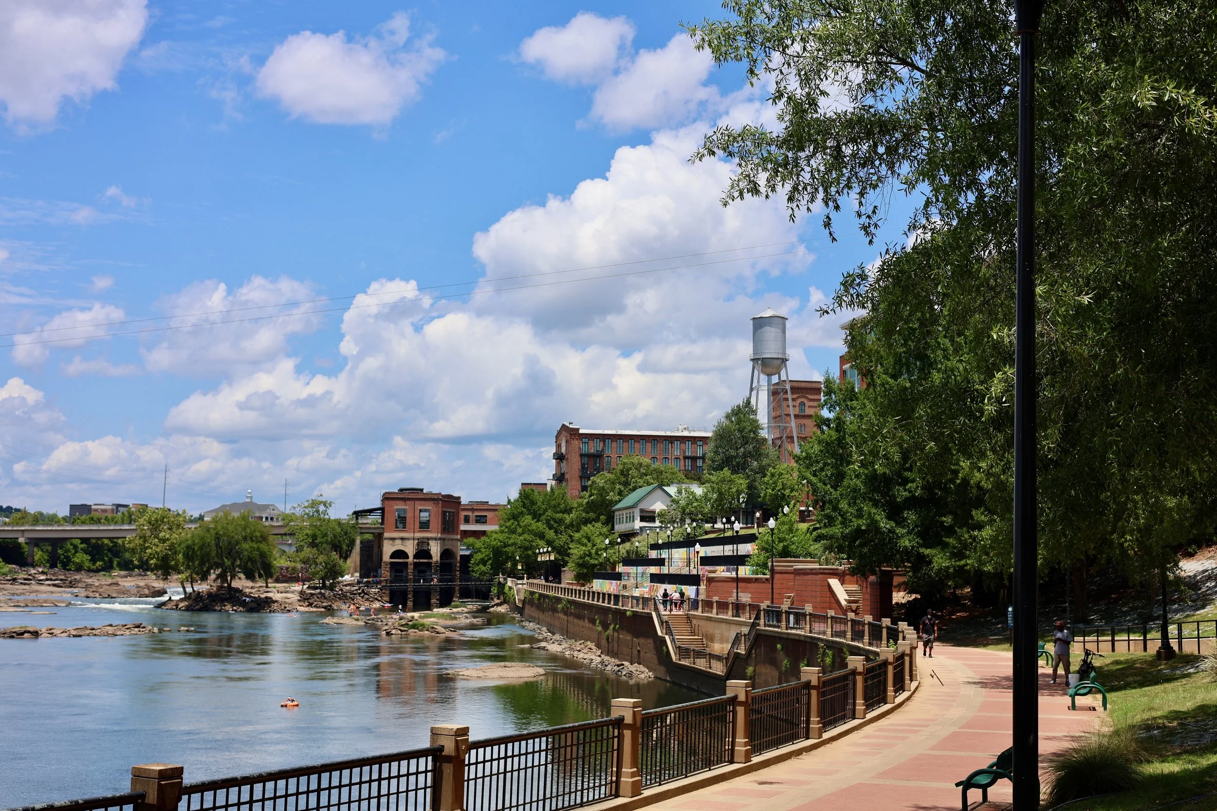 Riverwalk pathway with a concrete walkway, wooden railings, and several people walking along the river. Trees line the walkway, and buildings, including a water tower, are visible in the background under a partly cloudy sky.
