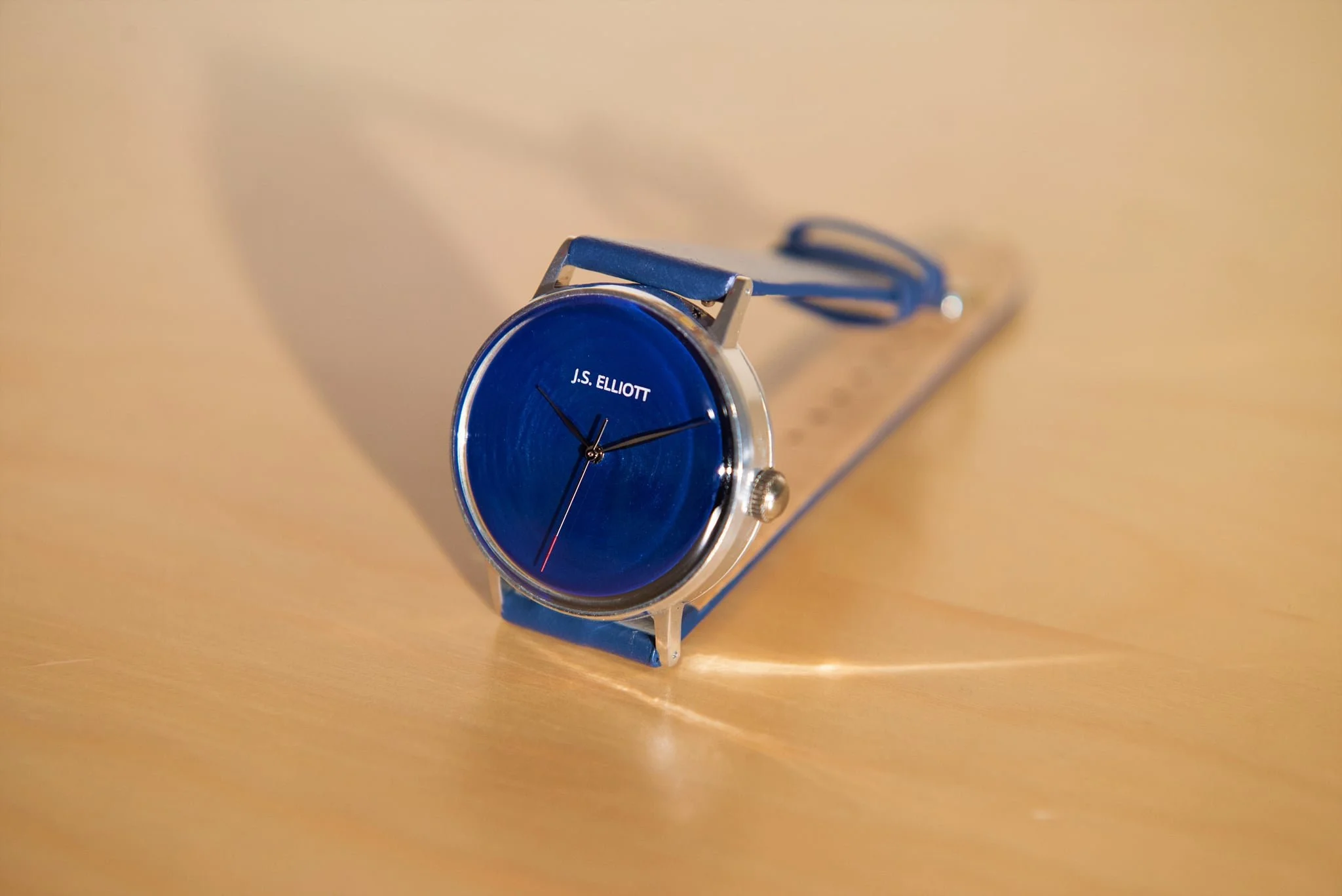 A blue wristwatch with the name JS Elliott on the dial, resting on a light wooden surface.